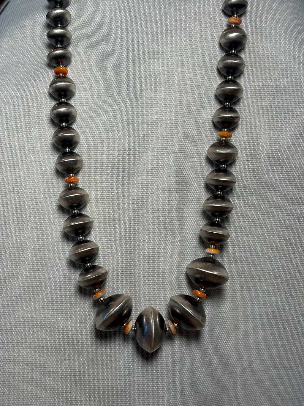 A beaded necklace with dark gray metallic disc beads and small orange spacer beads, displayed on a textured light gray fabric background.