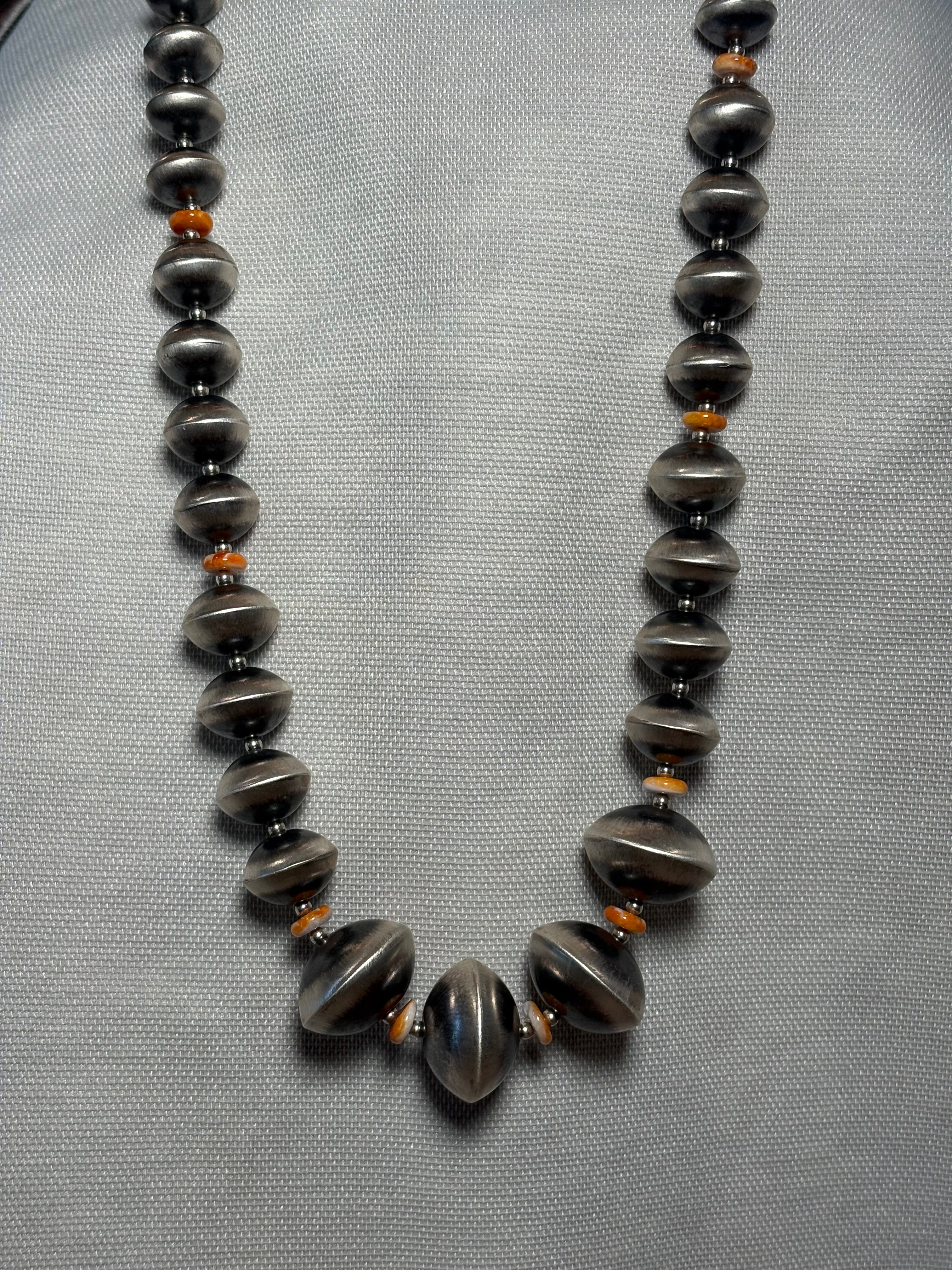 A beaded necklace with dark gray metallic disc beads and small orange spacer beads, displayed on a textured light gray fabric background.