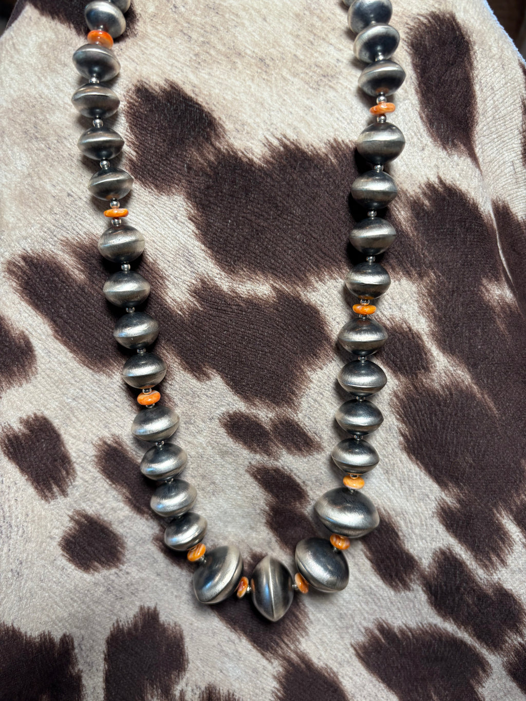 A beaded necklace with large, polished dark gray baroque-style beads and small orange spacer beads, displayed on a brown and beige animal-print faux fur surface.