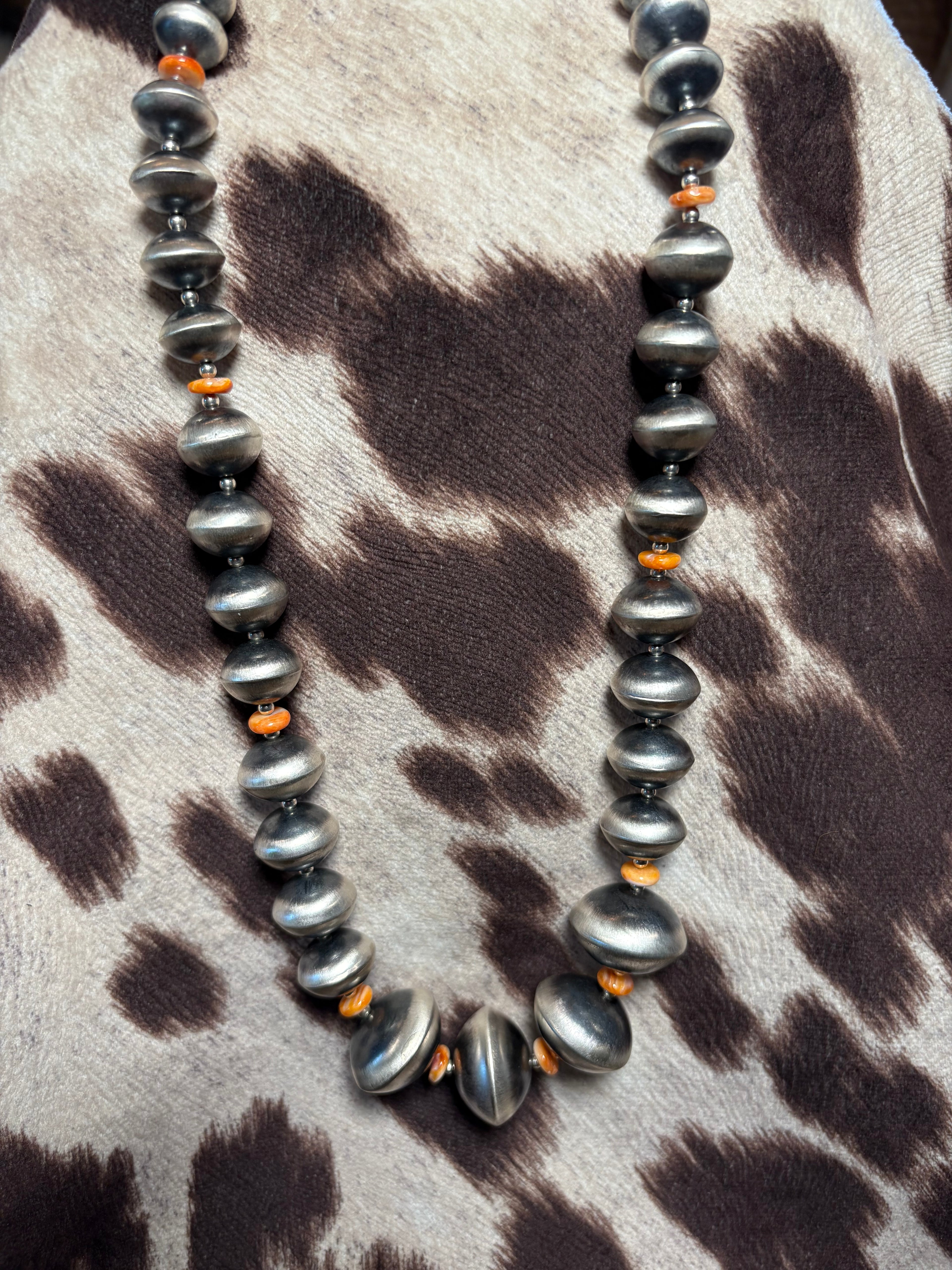 A beaded necklace with large, polished dark gray baroque-style beads and small orange spacer beads, displayed on a brown and beige animal-print faux fur surface.