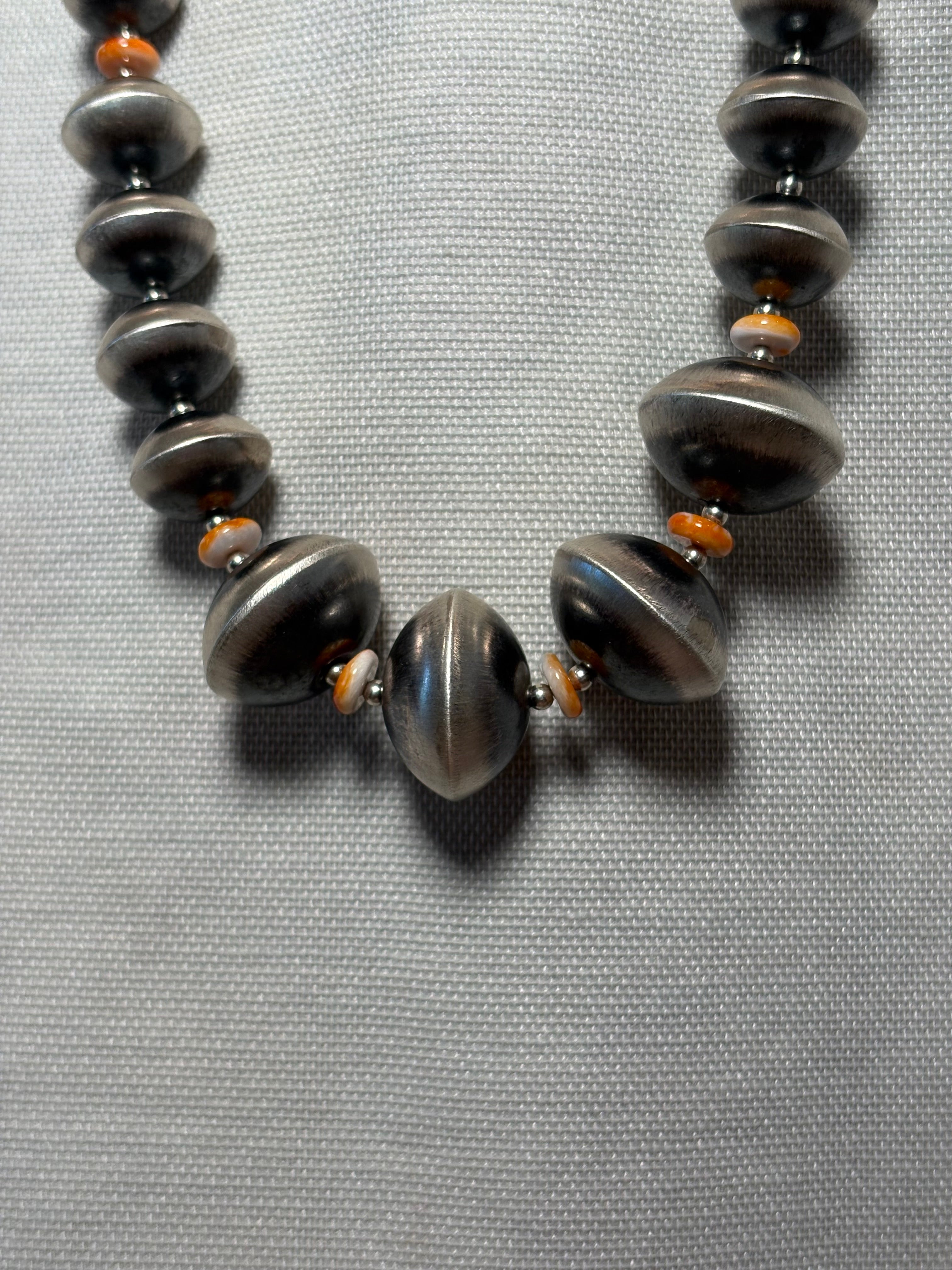 A close-up of a beaded necklace featuring large, polished dark gray metallic disc beads and smaller orange accent beads, laid flat on a textured gray fabric background.