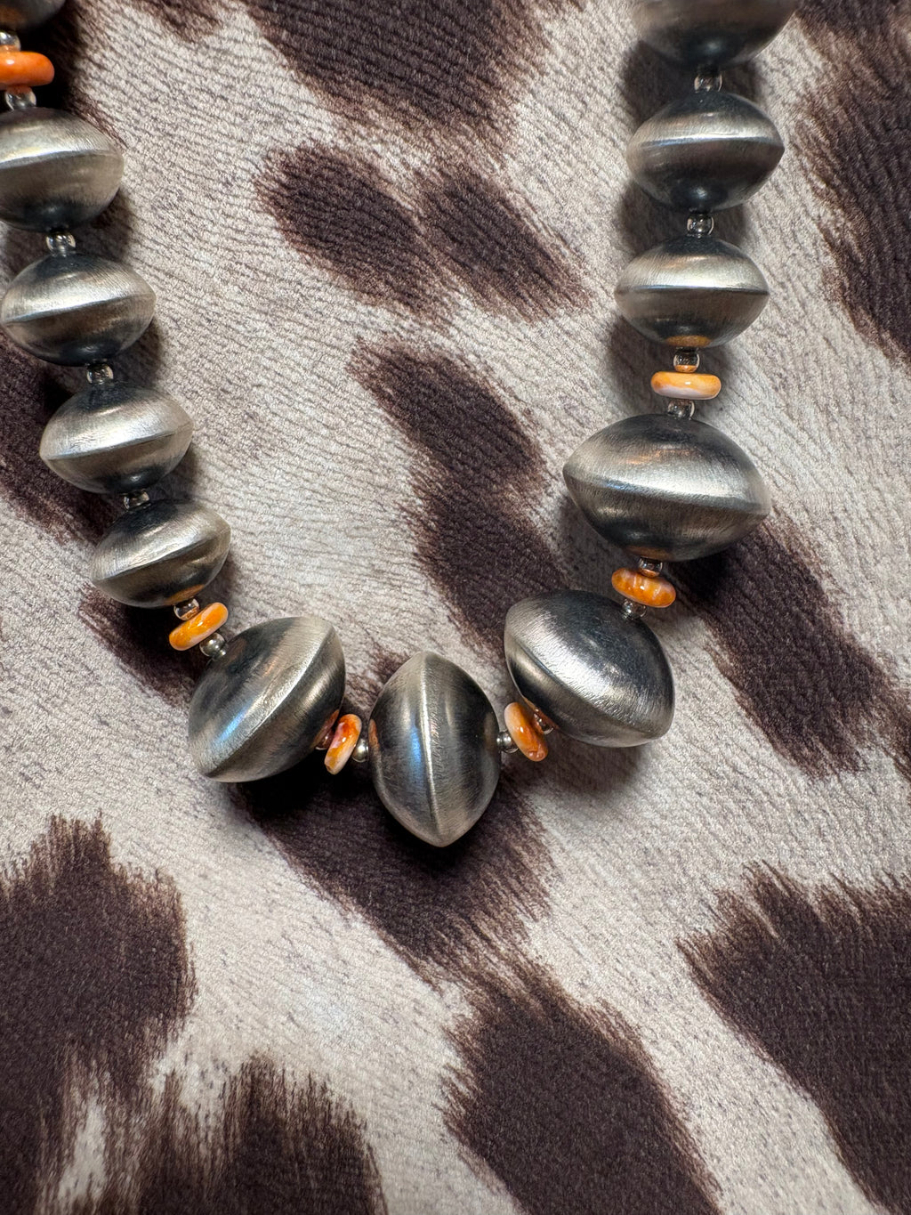 A close-up of a beaded necklace with large metallic silver-gray disc-like beads and small orange spacer beads, laid on a brown-and-cream animal-print fabric.