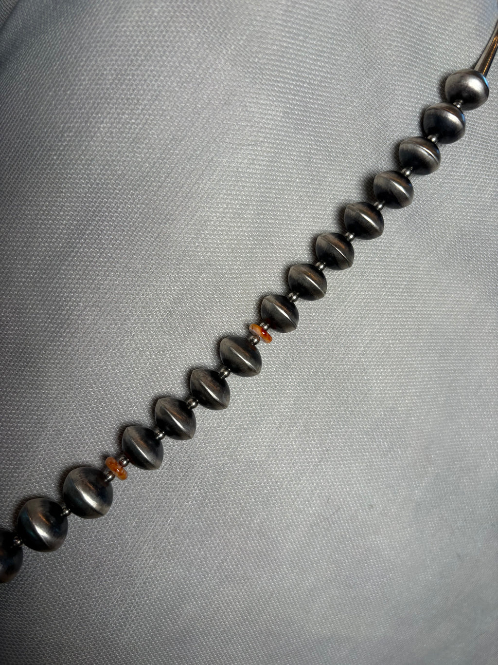 A close-up of a beaded strand with glossy dark metallic beads and a few small orange spacer beads, laid diagonally on a light gray textured fabric background.
