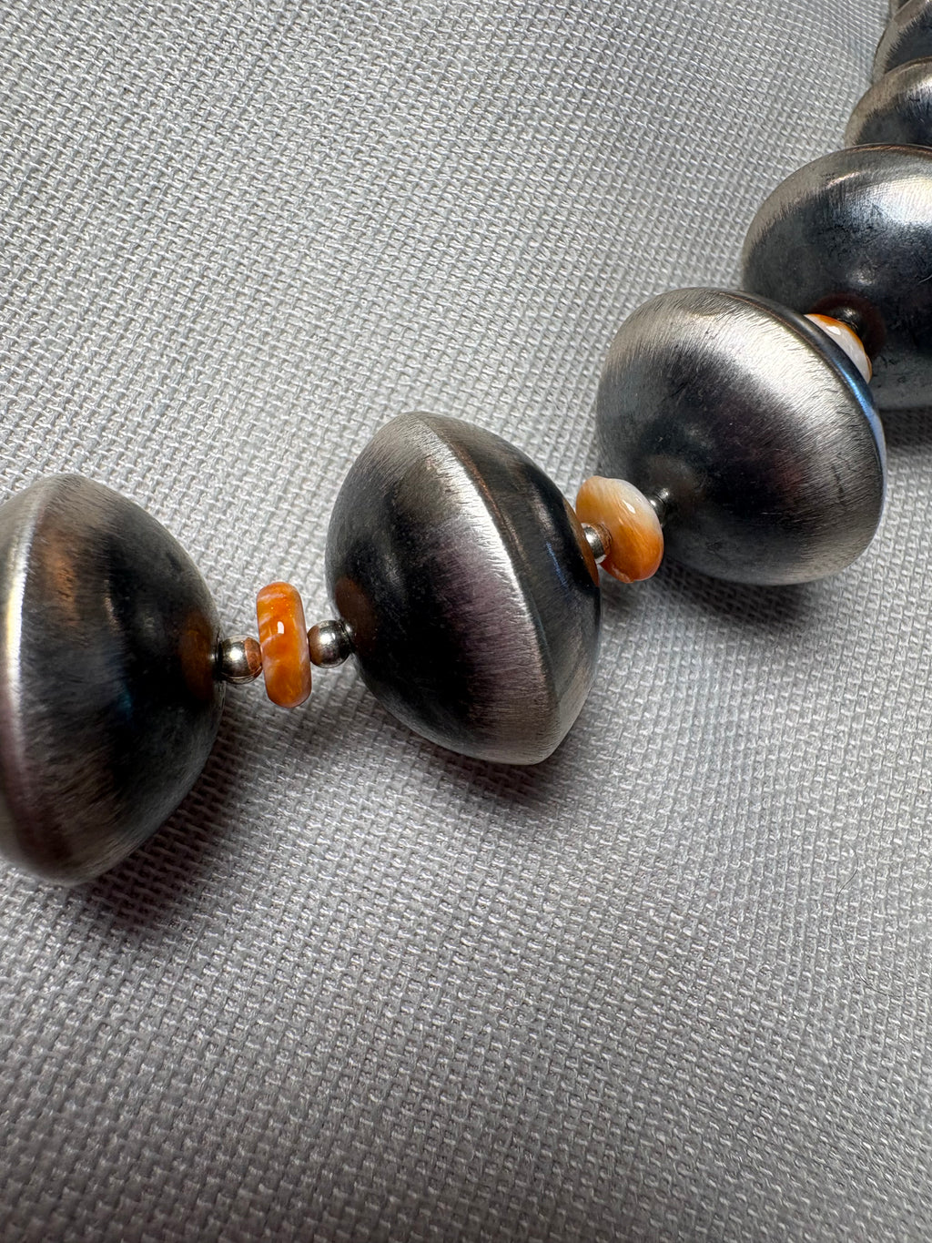 A close-up of a beaded necklace featuring large, dark metallic silver disc-like beads separated by small orange spacer beads, set against a textured gray background.