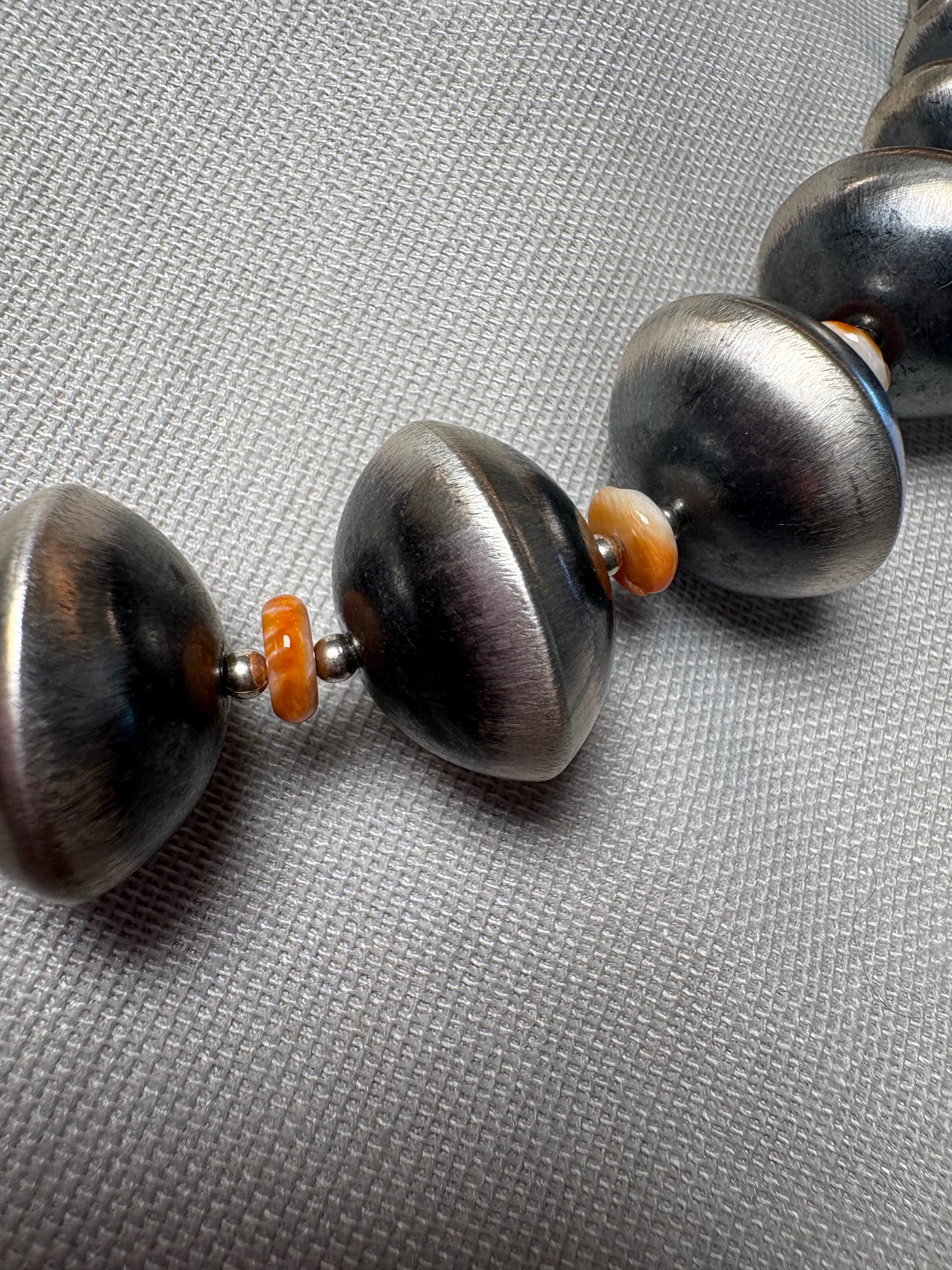 A close-up of a beaded necklace featuring large, dark metallic silver disc-like beads separated by small orange spacer beads, set against a textured gray background.