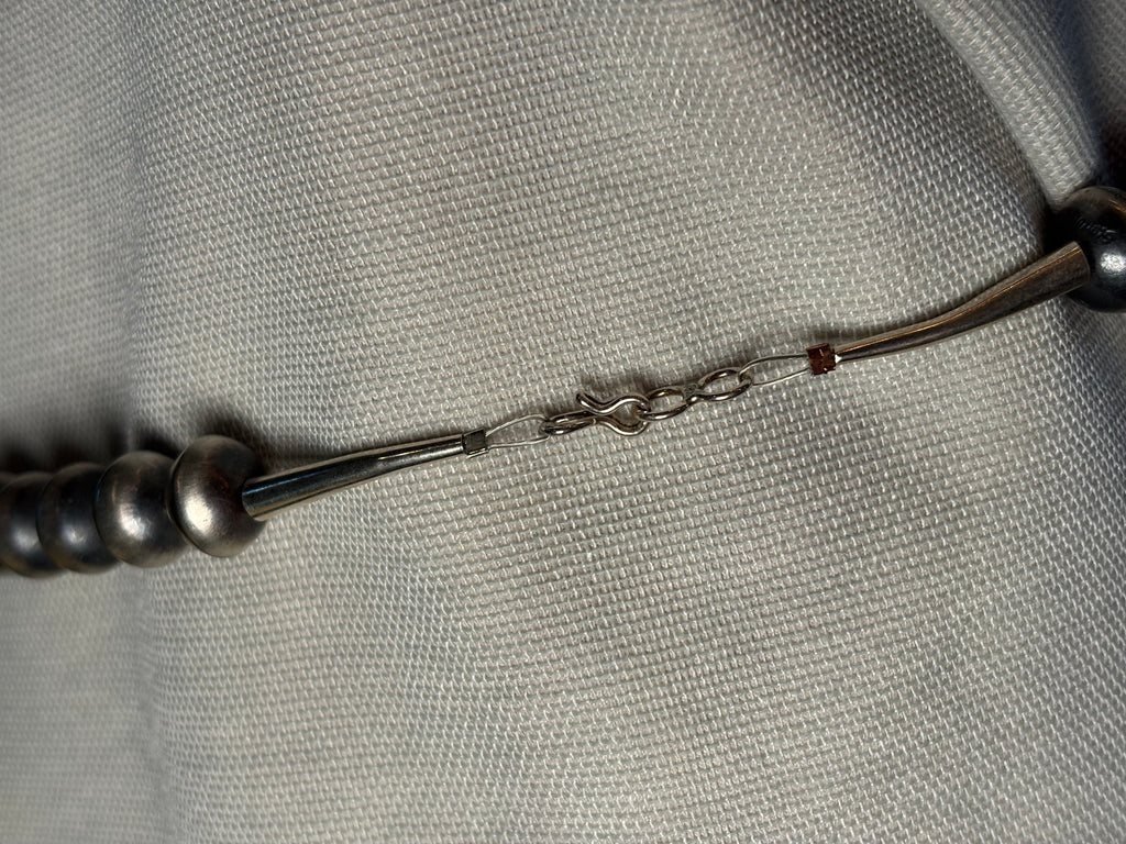 A close-up of a silver-toned beaded necklace or bracelet laid on a textured light gray fabric background, showing metallic beads, a chain connector, and a small red accent bead.