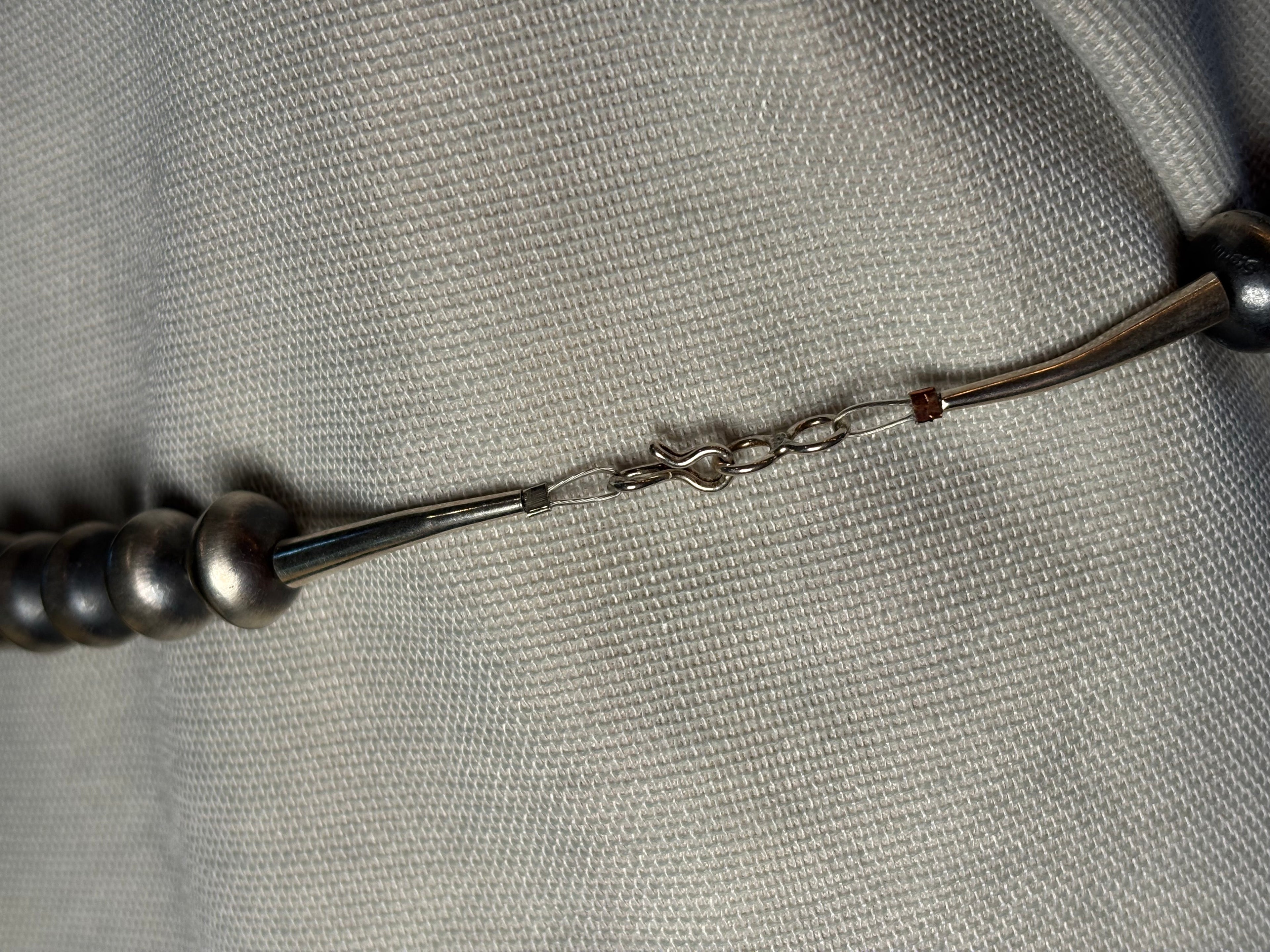 A close-up of a silver-toned beaded necklace or bracelet laid on a textured light gray fabric background, showing metallic beads, a chain connector, and a small red accent bead.