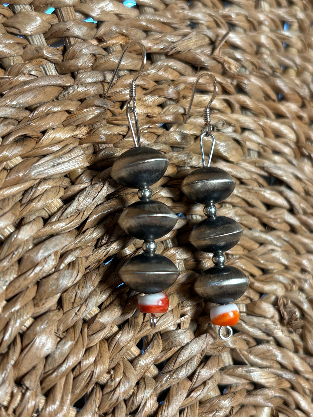 A pair of handmade dangle earrings with dark metallic beads and a small orange-and-white marbled bead, displayed on a woven brown wicker background.