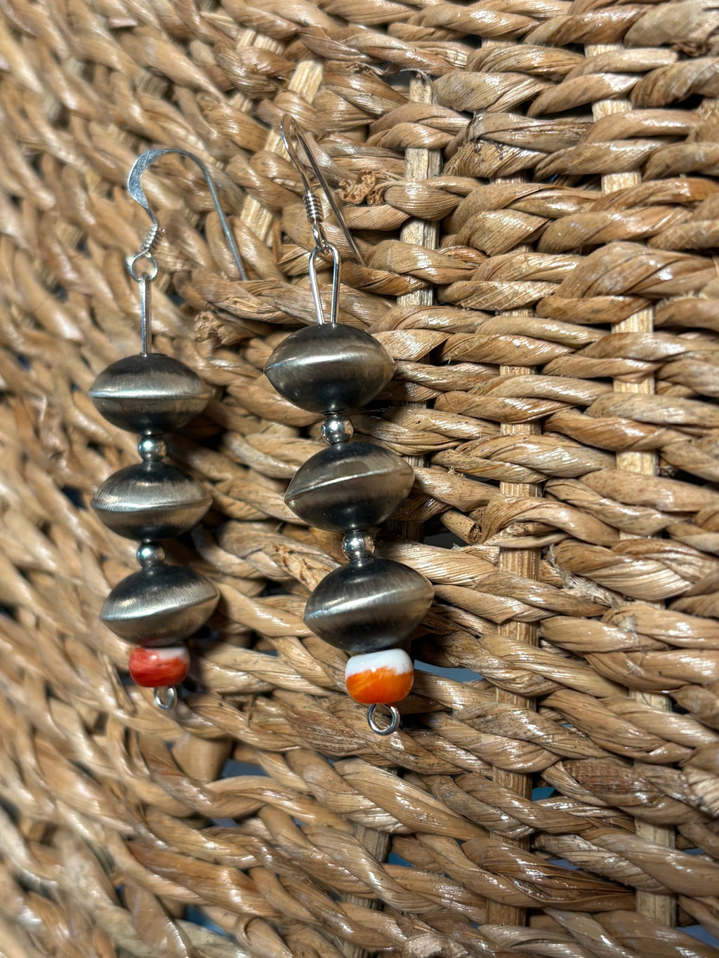 A pair of handmade dangle earrings with oxidized silver beads and small orange and white accent beads, displayed on a woven straw surface.