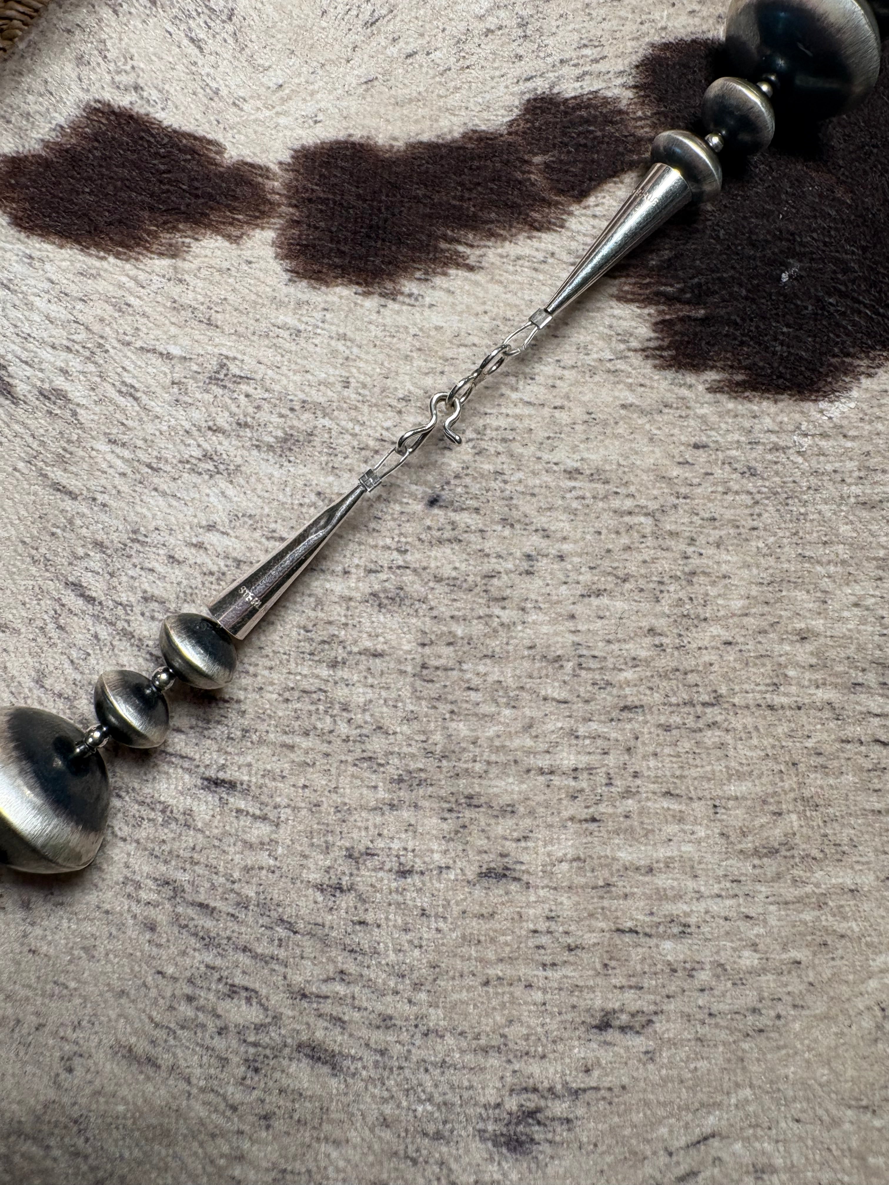 A close-up of a silver-toned metal tassel or decorative chain accessory with spherical beads, laid diagonally across a textured beige and gray surface with dark brown patches.
