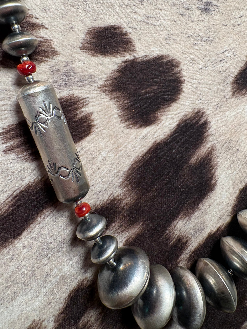 A close-up of a Southwestern-style beaded necklace featuring a large engraved silver cylinder bead, smaller silver disc beads, and red spacer beads, laid out on a brown and beige animal print rug.