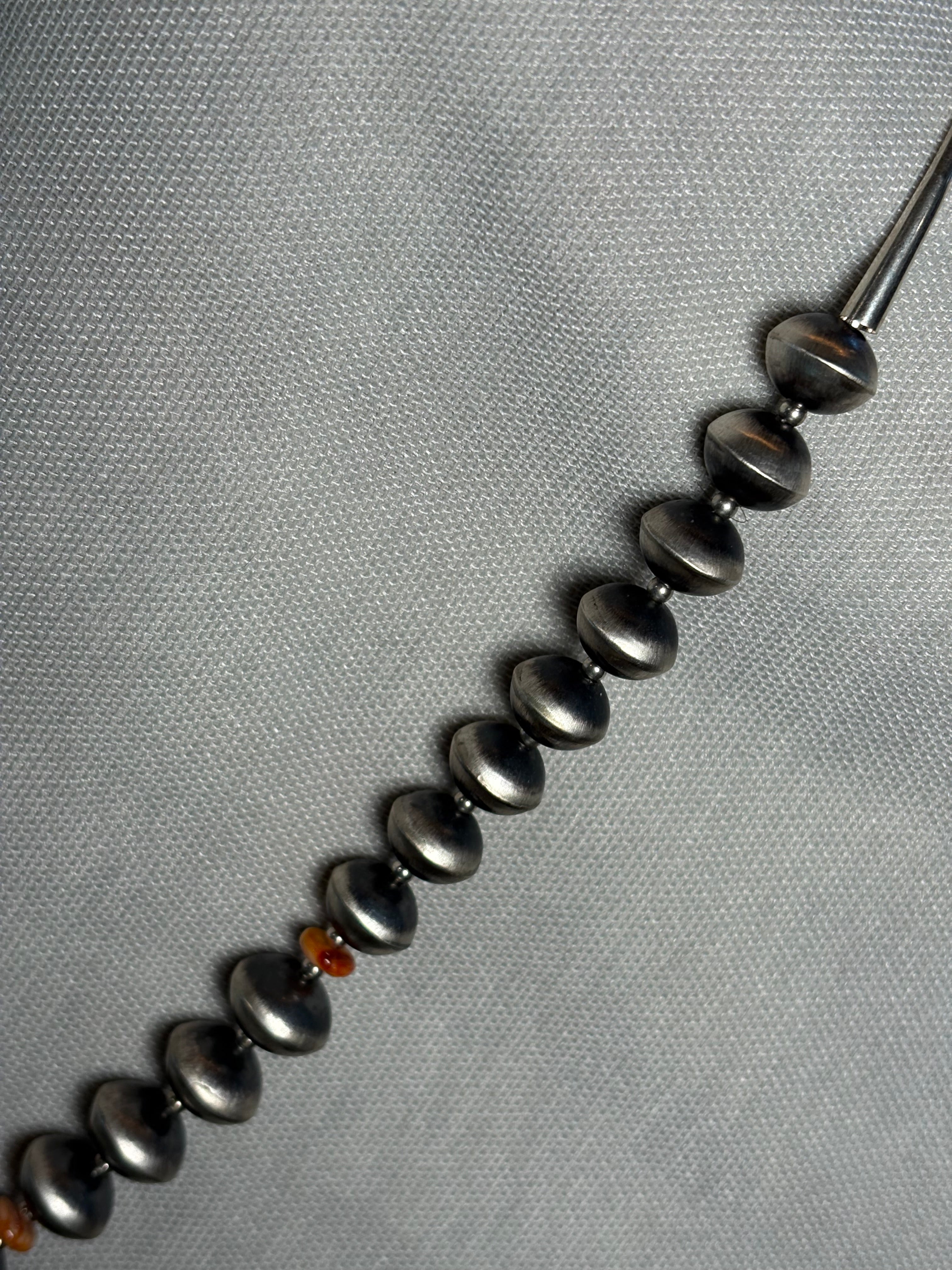 A close-up of a beaded necklace or bracelet laid diagonally on a textured gray fabric background. The beads are dark metallic gray with a slight iridescent sheen, and there are a few small warm-toned spacer beads near the bottom.