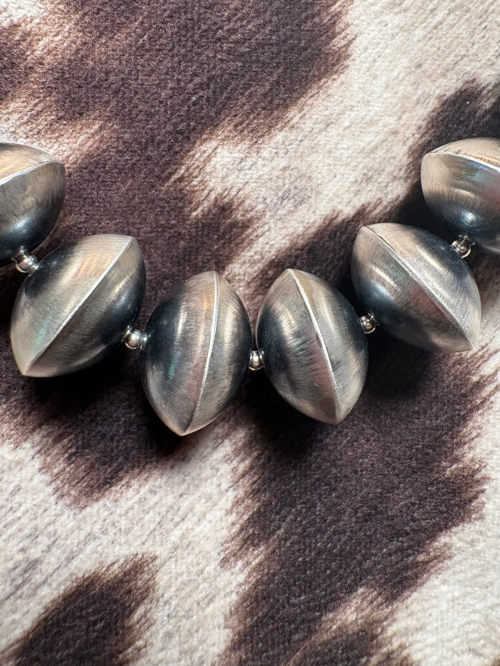 A close-up of a chunky beaded necklace with metallic gunmetal gray, football-shaped beads resting on a brown and cream cow-print fabric.