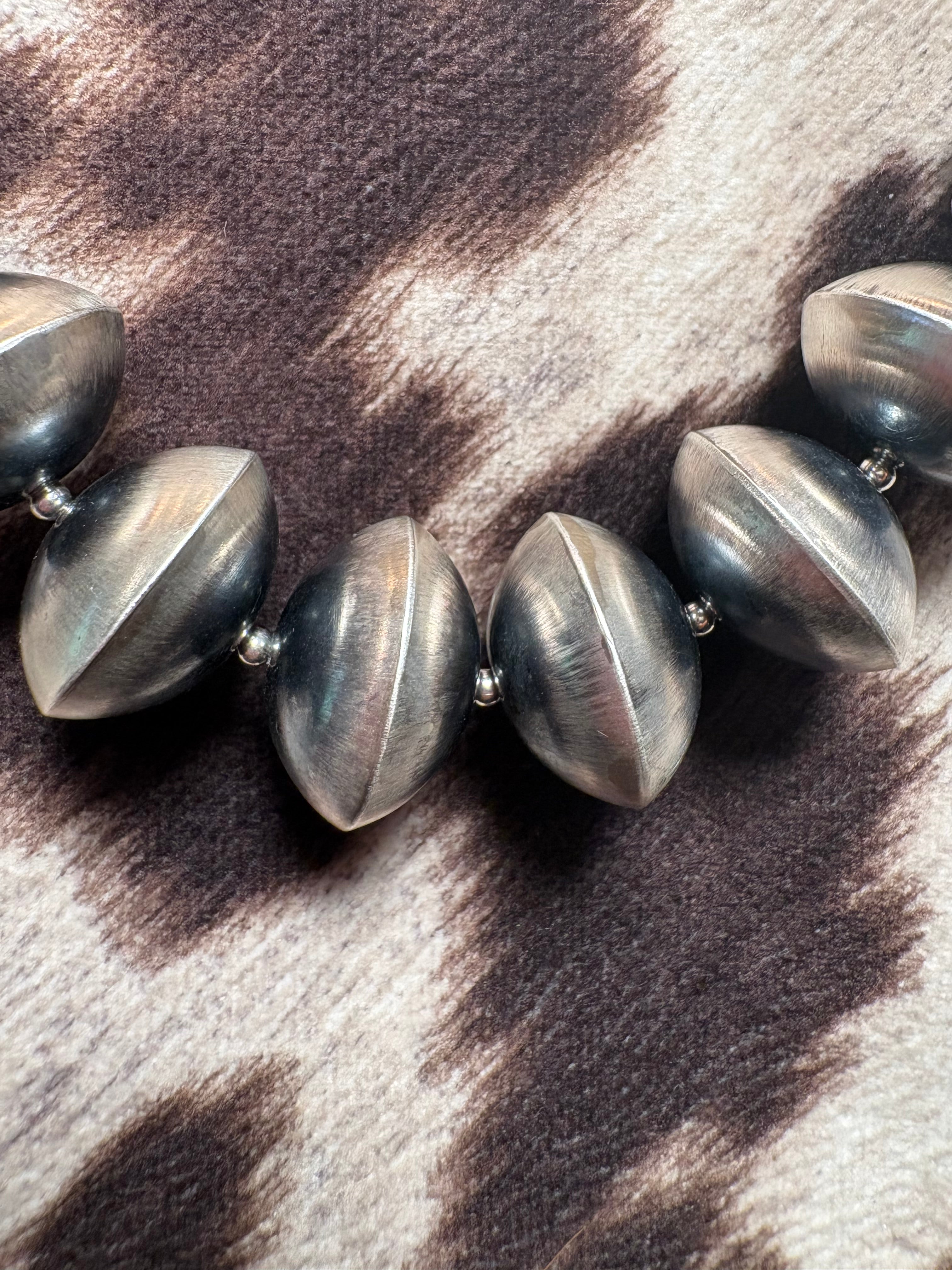 A close-up of a chunky beaded necklace with metallic gunmetal gray, football-shaped beads resting on a brown and cream cow-print fabric.