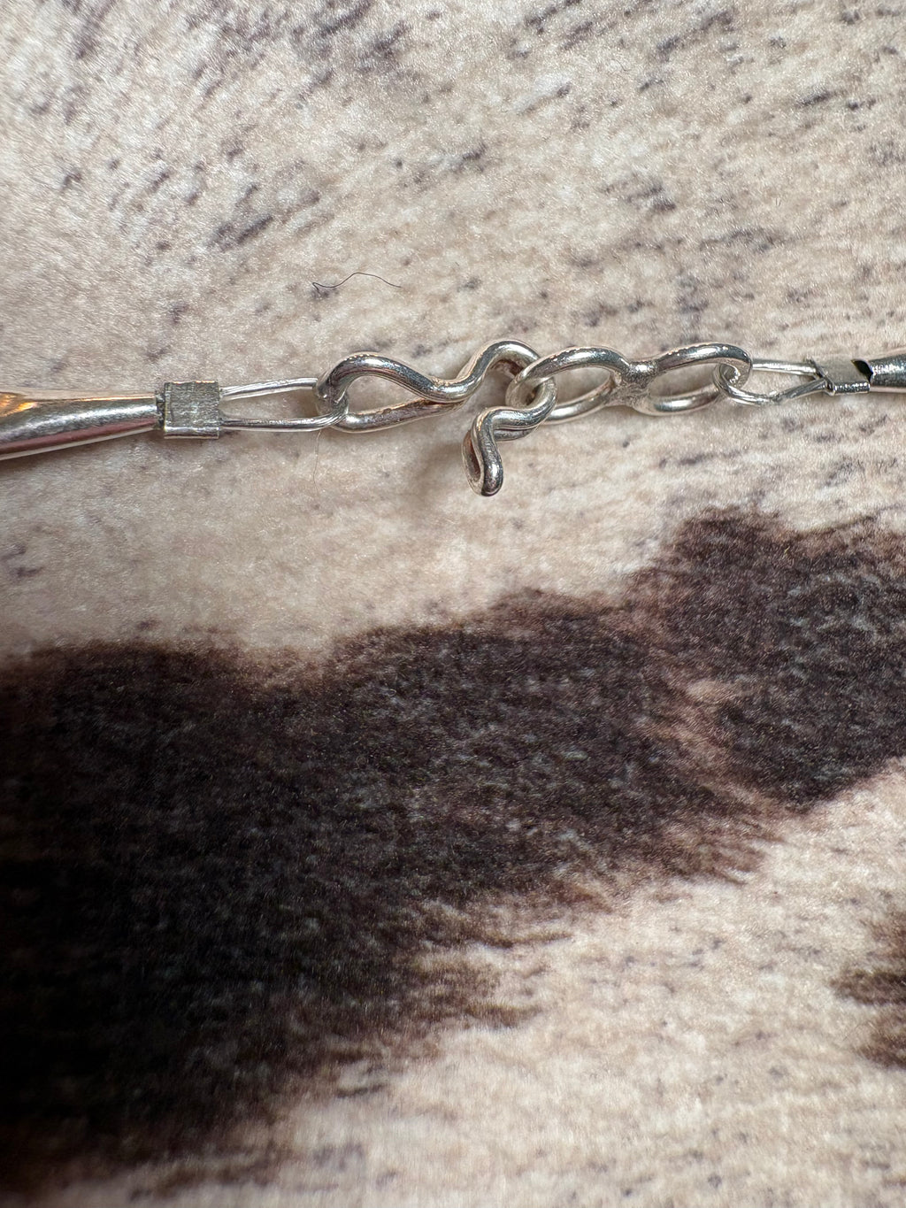 Close-up of a silver-toned metal chain bracelet or necklace clasp resting on a textured cowhide surface with dark brown and cream patches.