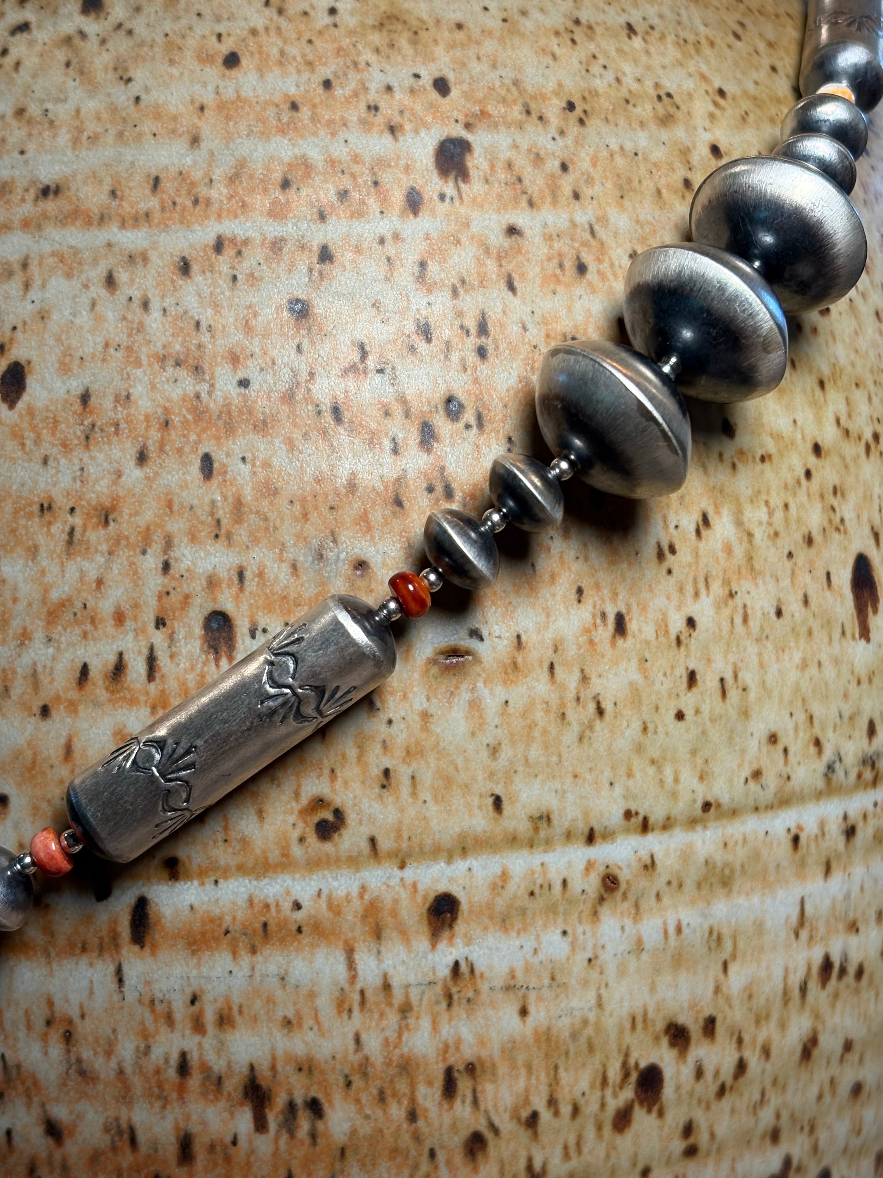 A close-up of a beaded necklace featuring oxidized silver-toned beads, a carved cylindrical centerpiece, and small red spacer beads, laid diagonally on a textured brown ceramic surface.
