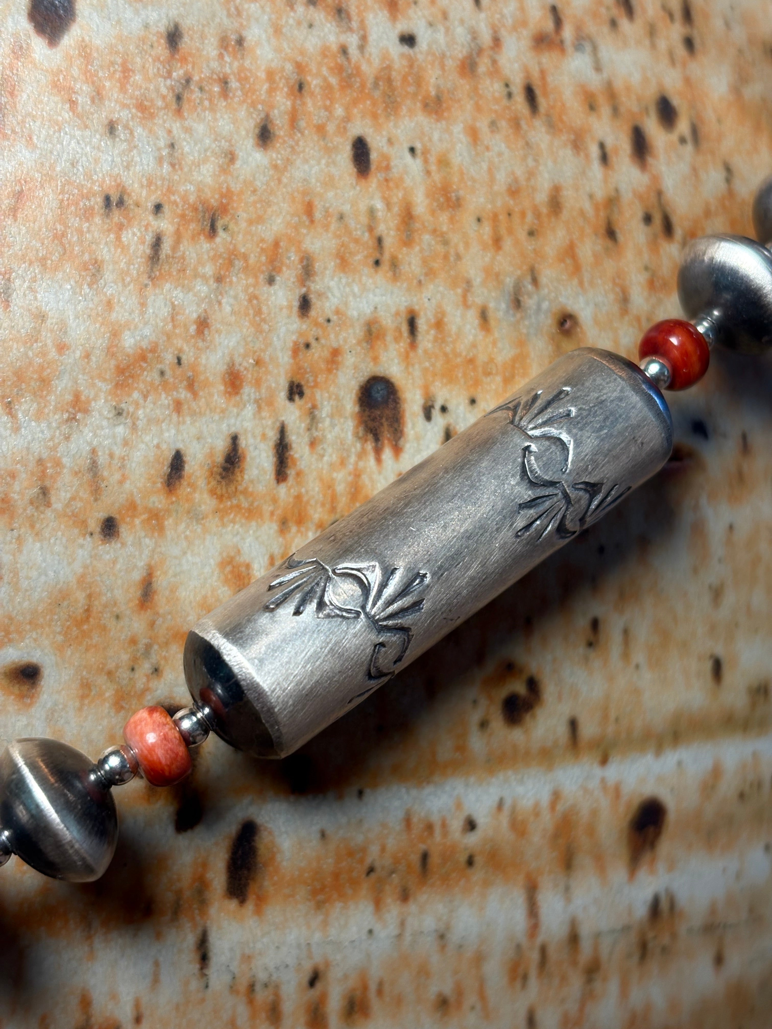 A close-up of a beaded necklace featuring a central cylindrical silver bead with engraved floral patterns, flanked by small red beads and metallic silver spacer beads, set against a textured rusted metal background.