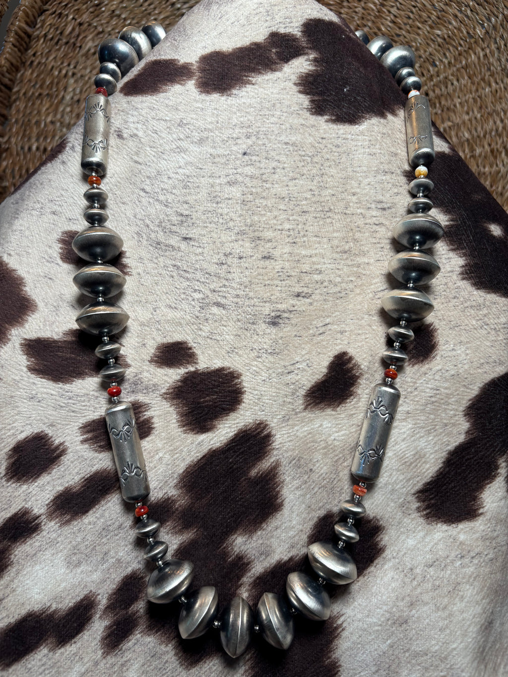 A handmade beaded necklace featuring distressed silver-toned metal discs, polished silver-toned cylindrical beads, and small red accent beads, displayed on a cowhide rug.