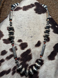 A handmade beaded necklace featuring distressed silver-toned metal discs, polished silver-toned cylindrical beads, and small red accent beads, displayed on a cowhide rug.