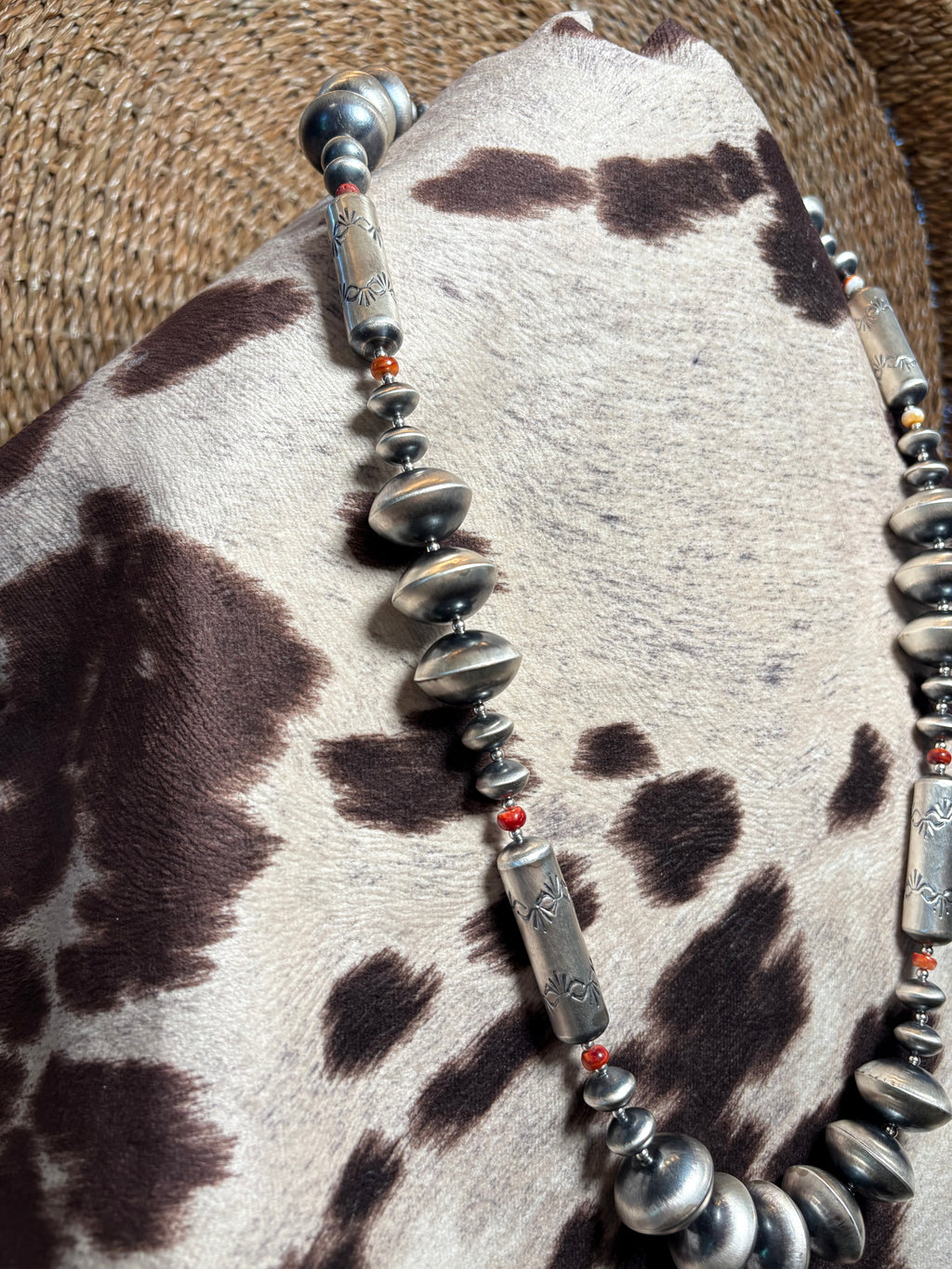 A close-up of a western-style beaded necklace with silver-toned disc and cylindrical beads, small red spacer beads, and dark metallic accents, draped over a cowhide surface.