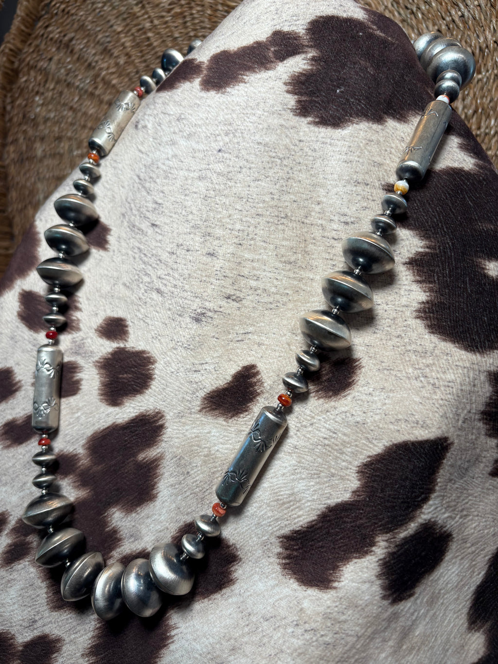 A beaded necklace with metallic silver-toned disc and cylindrical beads, accented by small red beads, displayed on a cowhide surface.