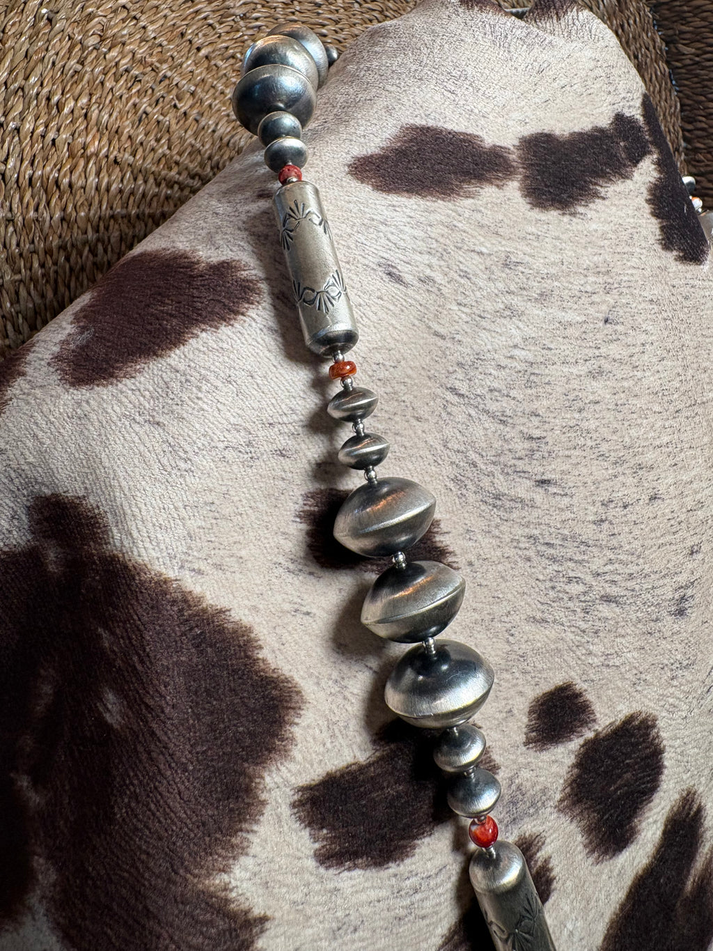 A close-up of a silver-toned beaded necklace or bracelet laid diagonally across a cowhide surface, with small red beads and engraved cylindrical accents.