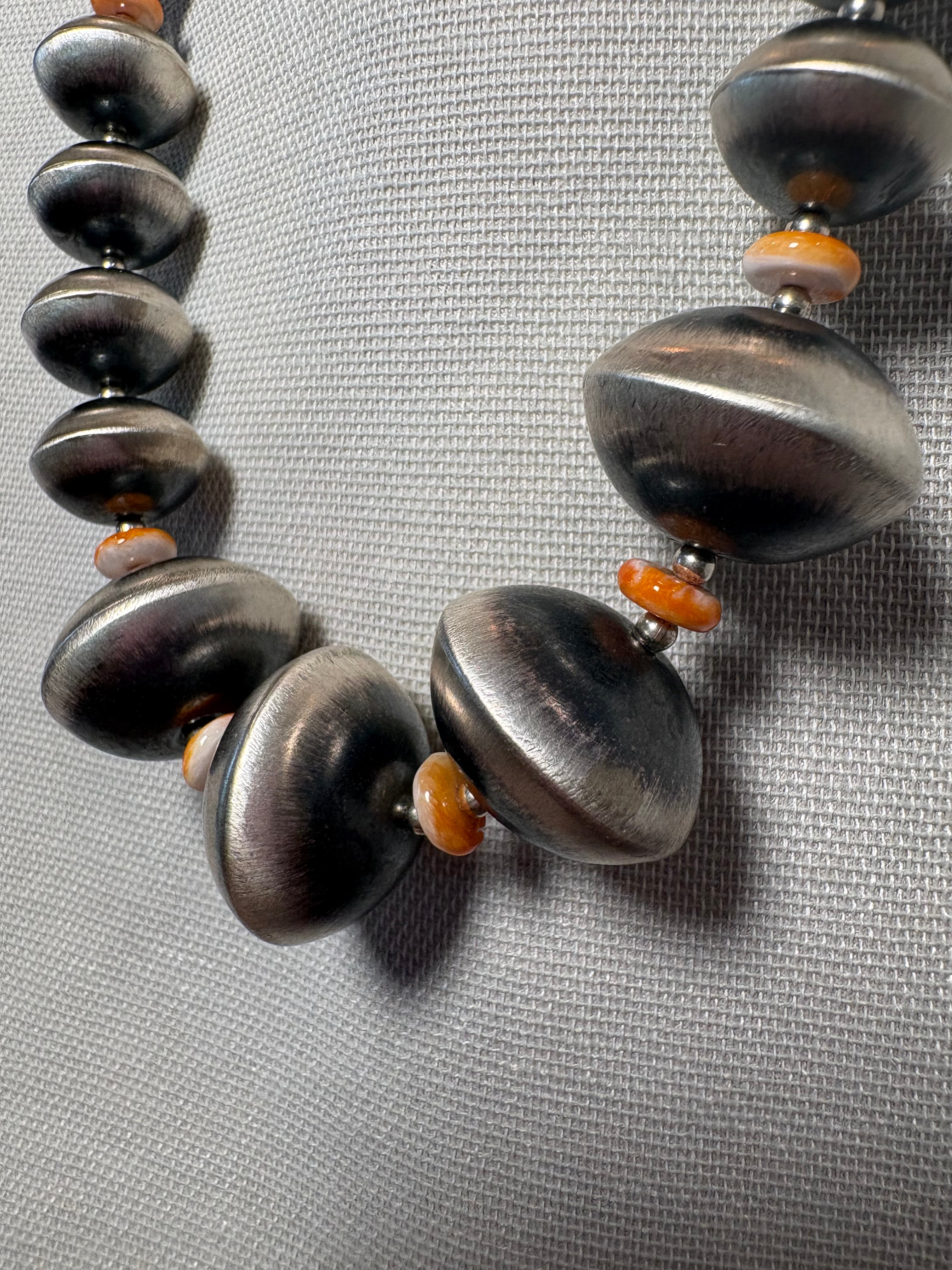 A close-up of a beaded necklace with large, dark metallic gray disc-shaped beads and small orange spacer beads, displayed on a textured gray background.