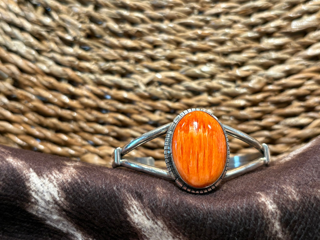 A silver-toned split-band ring featuring a large, oval cabochon orange stone with visible wood grain, resting on a dark brown and white cowhide surface with a woven straw background.