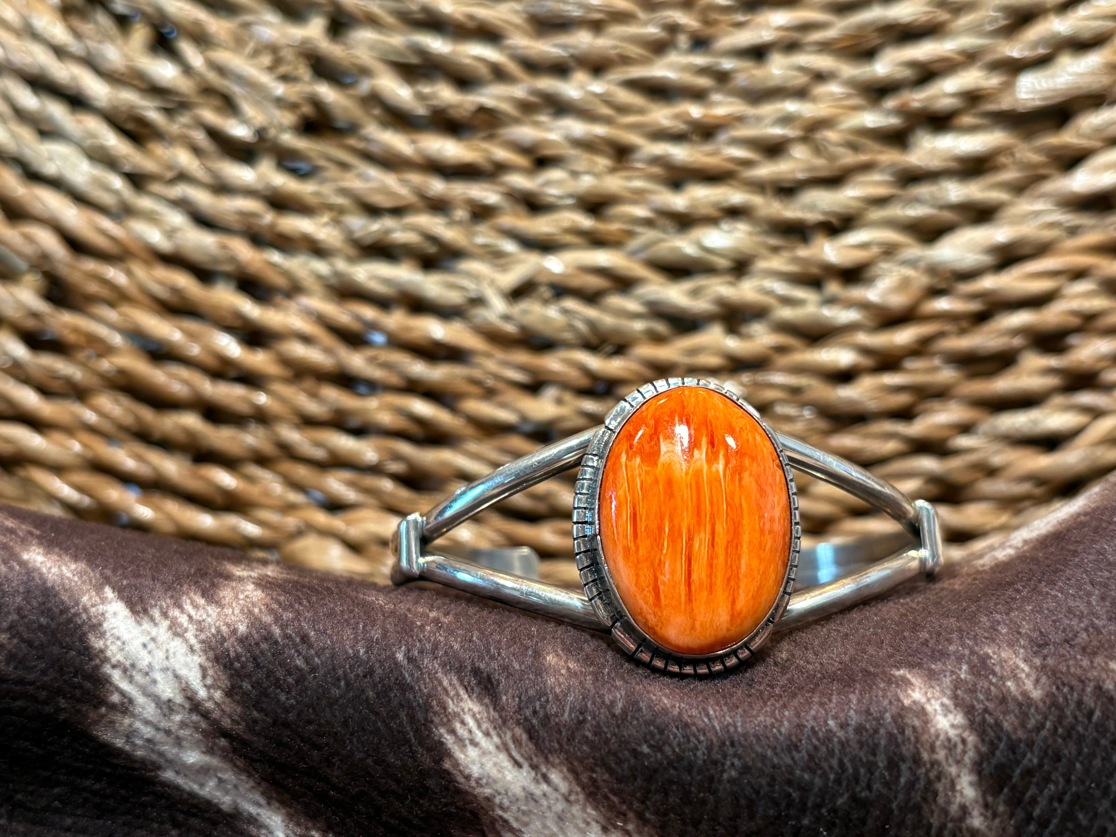 A silver-toned split-band ring featuring a large, oval cabochon orange stone with visible wood grain, resting on a dark brown and white cowhide surface with a woven straw background.
