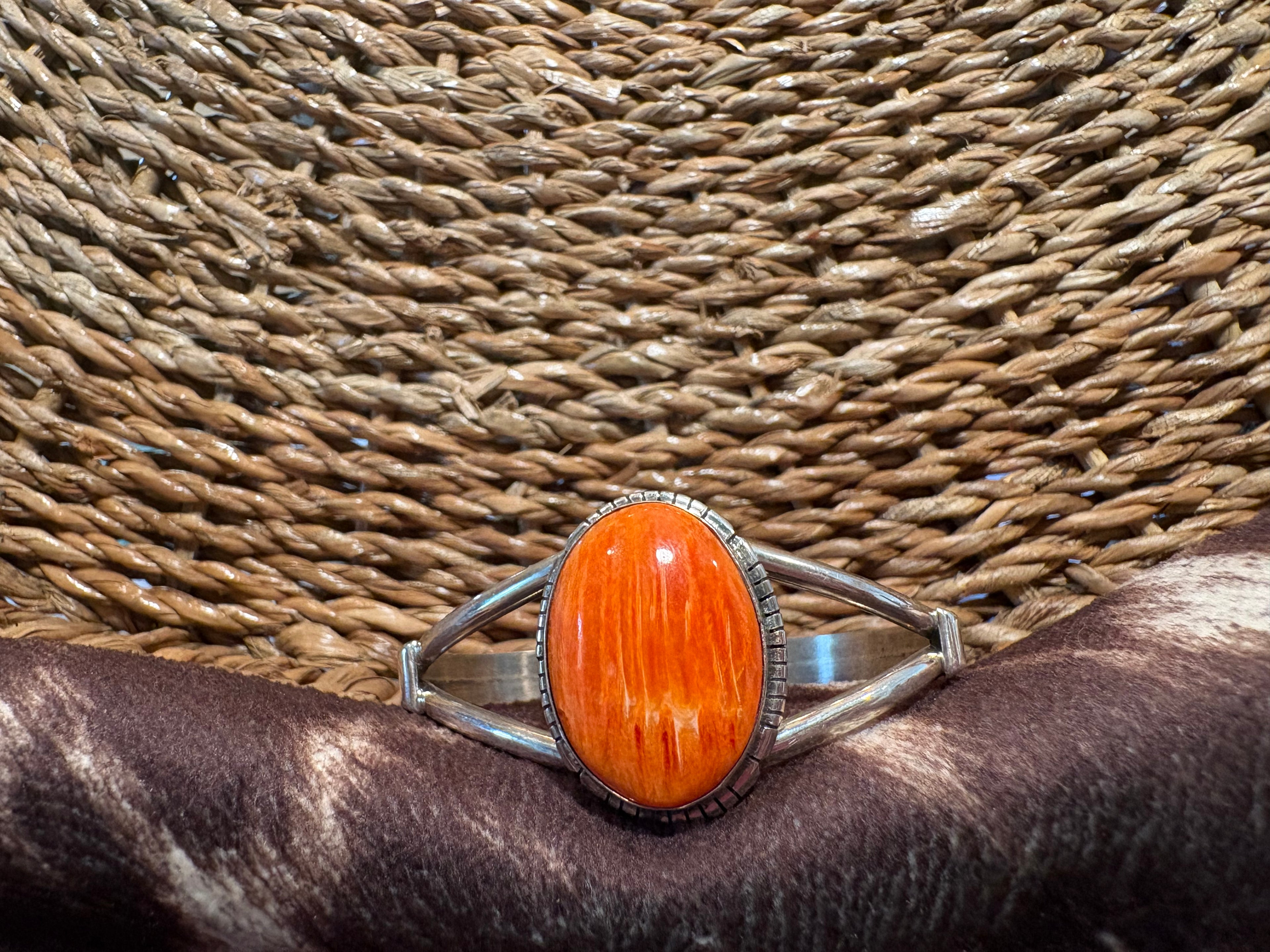 A silver-toned cuff ring featuring a large oval cabochon of vibrant orange woodgrain stone, displayed on a dark brown fur surface against a woven straw background.