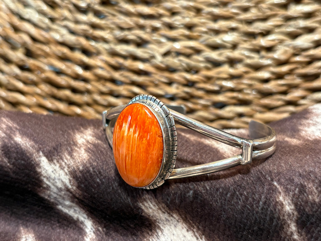 A sterling silver cuff ring featuring a large, oval cabochon orange stone with visible wood grain, resting on a dark brown and white zebra-patterned surface with a woven straw background.