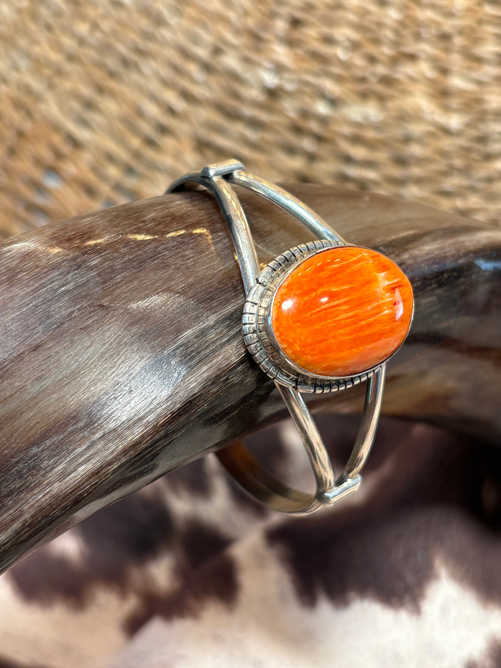 A silver-toned wrap-style ring with a large oval orange cabochon stone, displayed on a dark brown horn or antler against a textured tan and brown background.