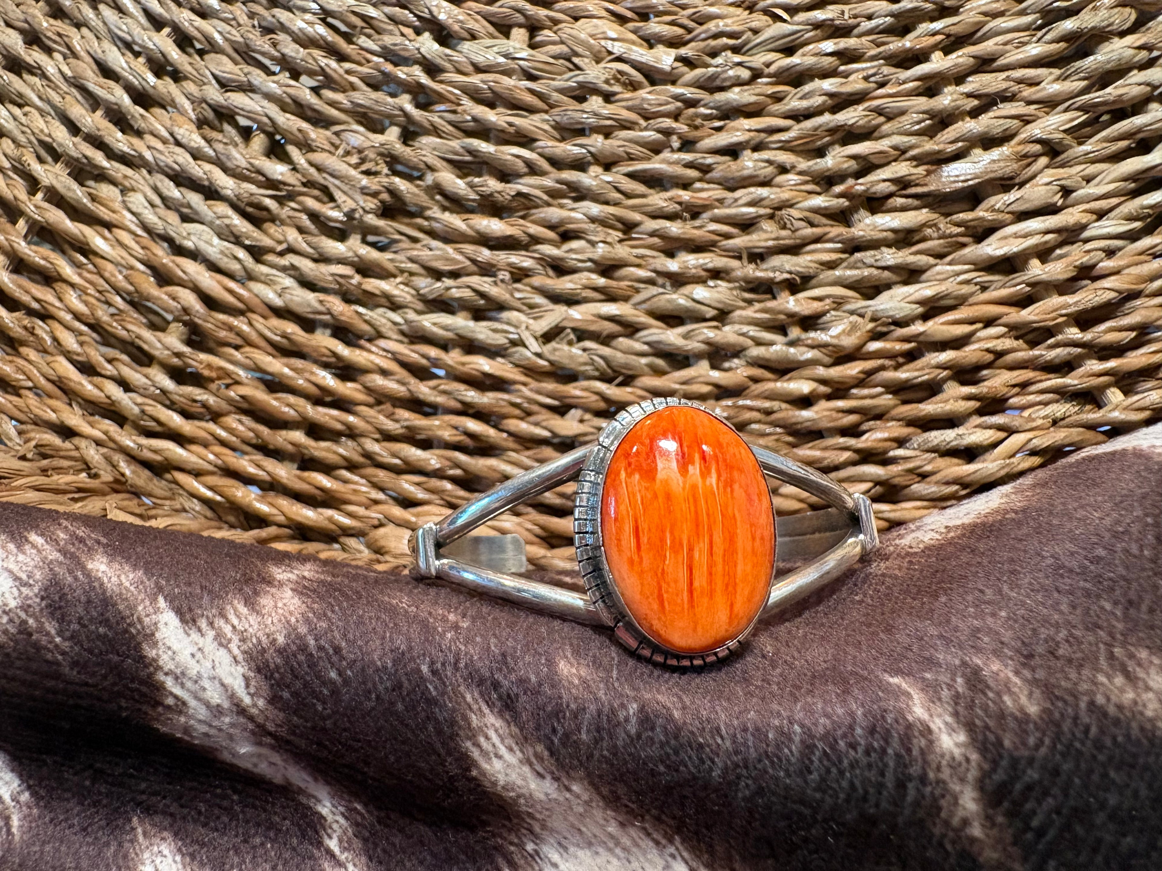 A silver cuff ring featuring a large, polished orange oval cabochon stone, displayed on a dark brown and white zebra-patterned hide against a woven straw background.