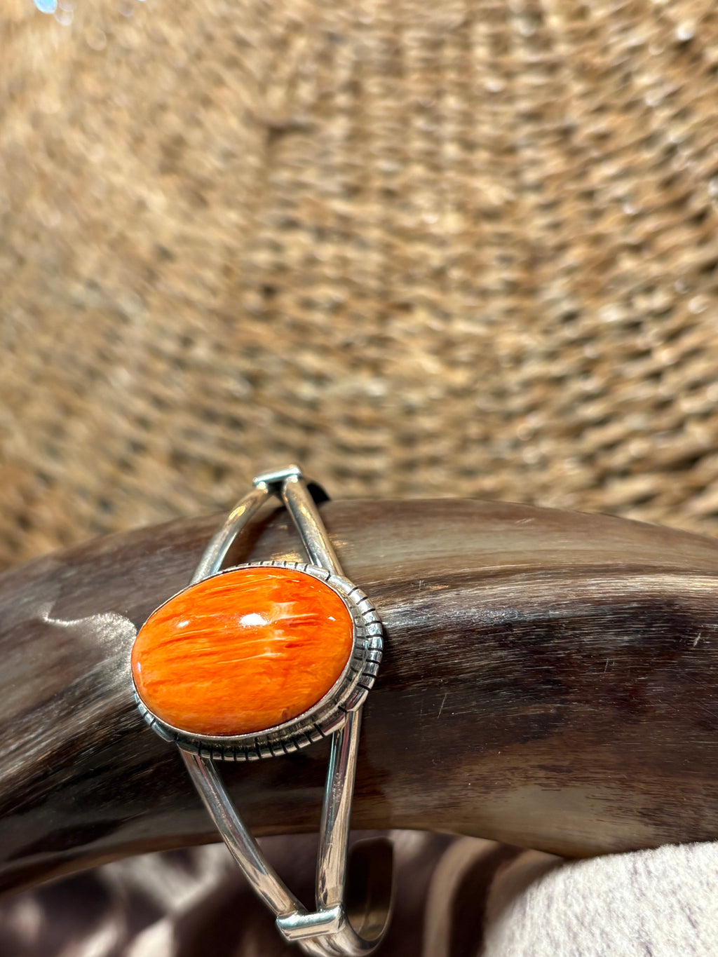A silver-toned cuff bracelet featuring a large, polished orange cabochon gemstone, displayed on a dark wooden stand against a woven straw background.