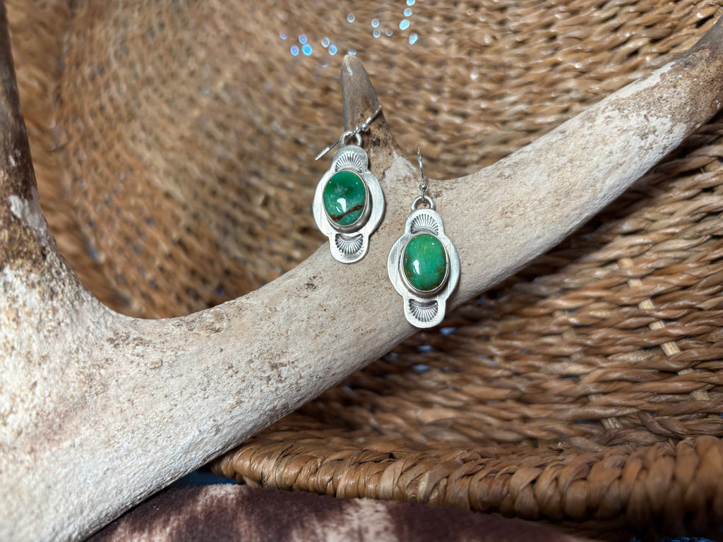 A pair of silver dangle earrings with oval green gemstones, displayed on a light-colored antler over a woven brown basket.