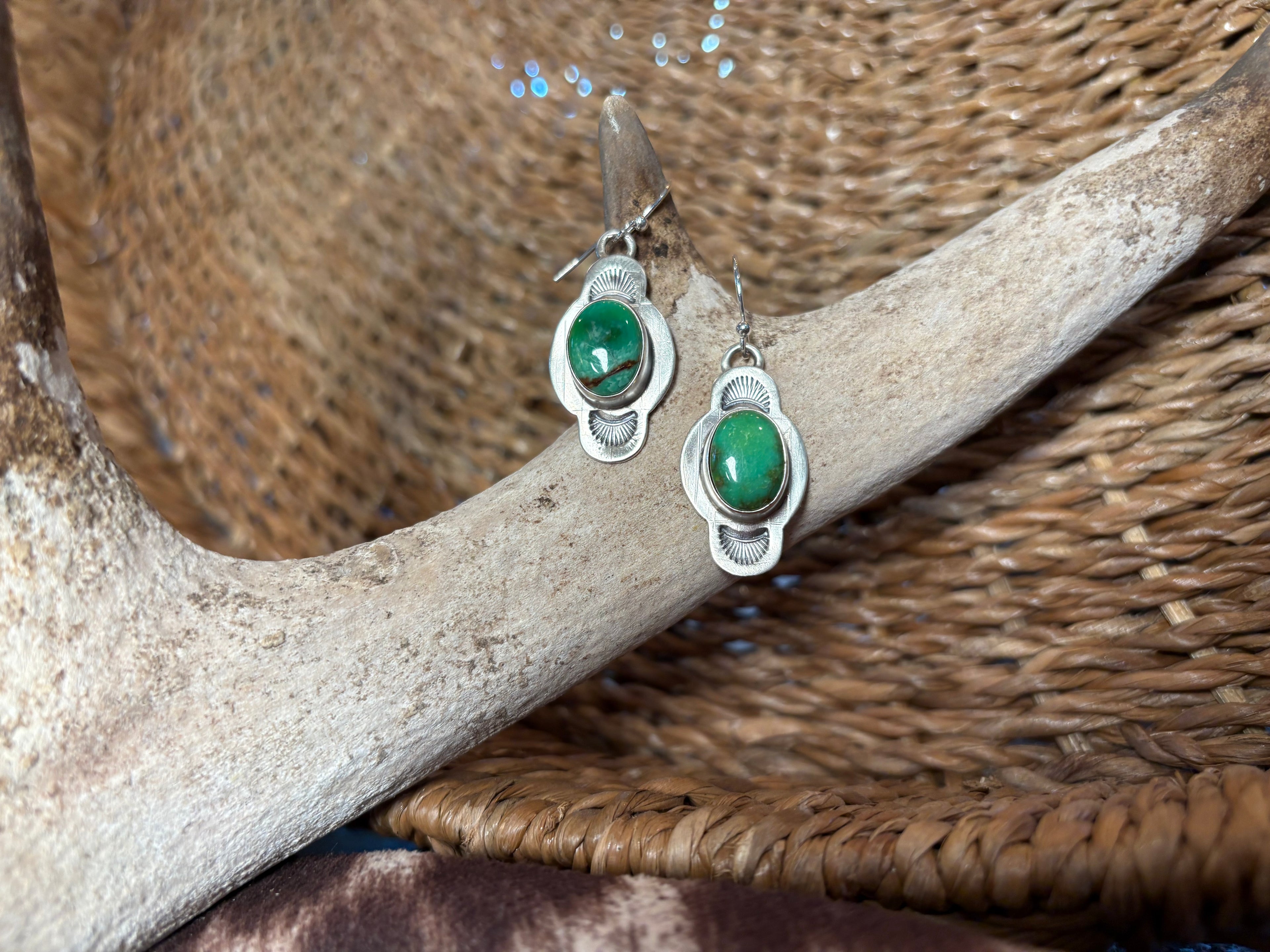 A pair of silver dangle earrings with oval green gemstones, displayed on a light-colored antler over a woven brown basket.