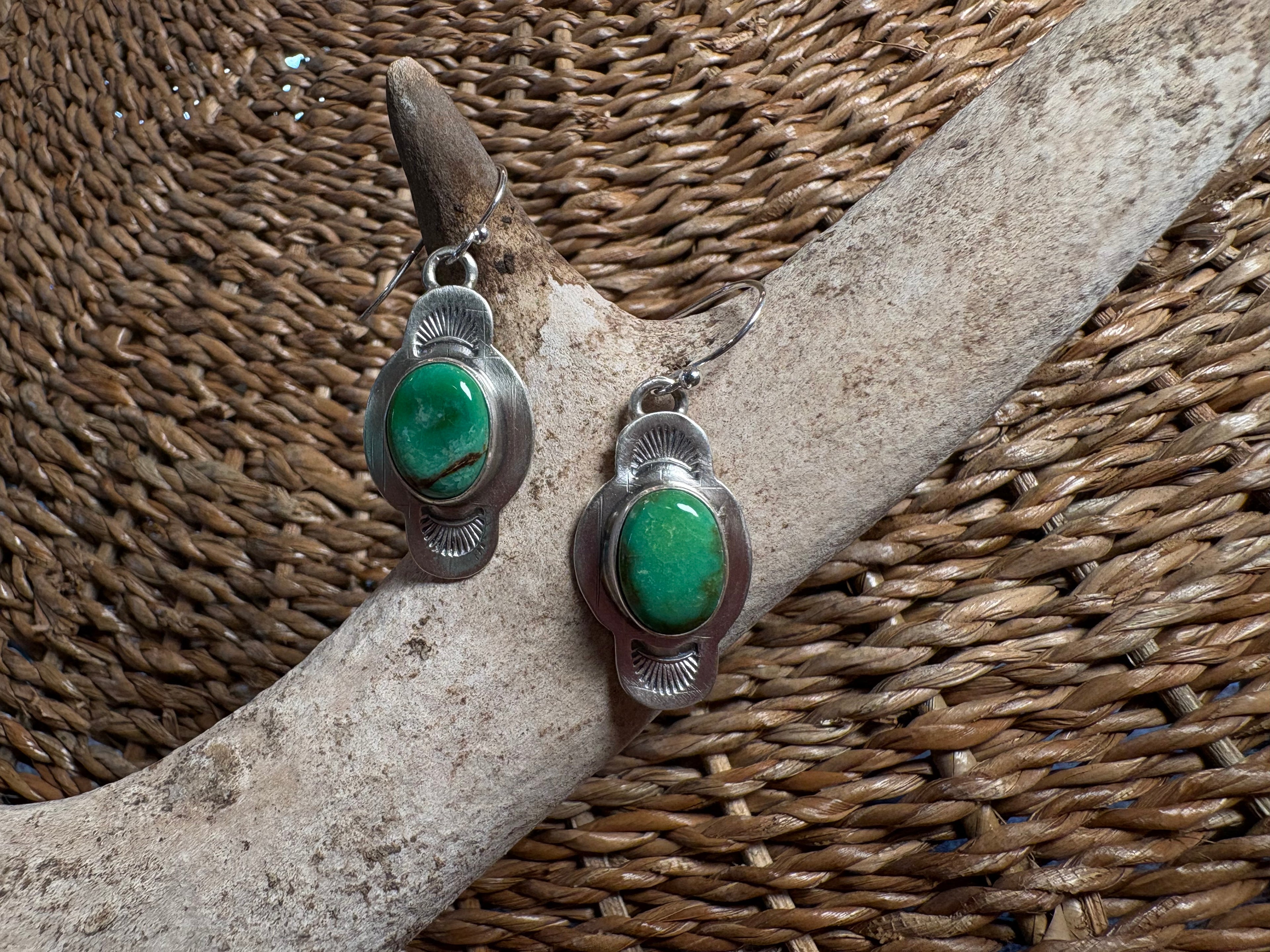 A pair of silver-toned dangle earrings with oval green stone centers, displayed on a light wood branch over a woven brown basket background.