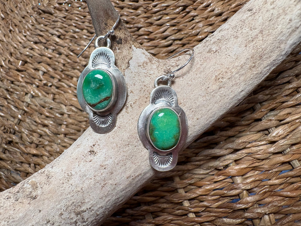 A pair of silver dangle earrings with oval green stone centers, displayed on a light wood branch over a woven brown basket background.