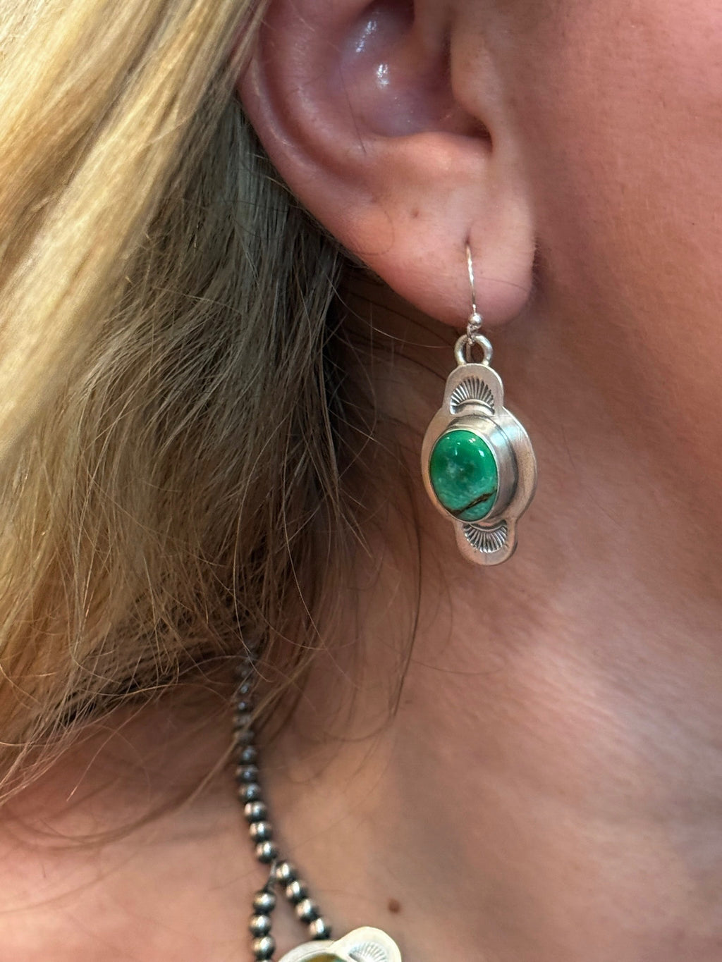 A close-up of a woman's ear wearing a silver-toned dangle earring with a round green stone, next to her blonde hair and a dark beaded necklace.