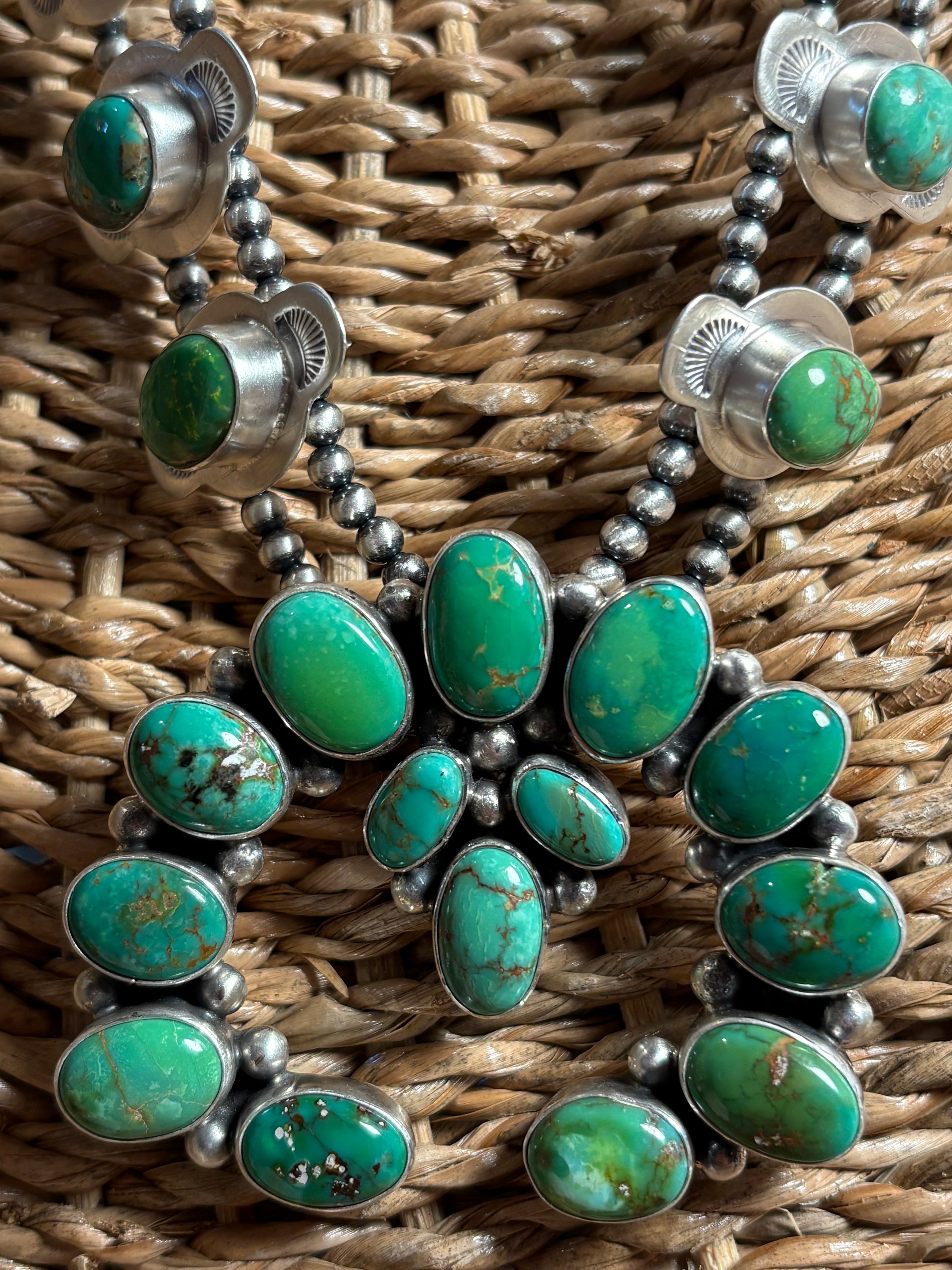 A close-up of a silver and turquoise necklace laid flat on a woven brown wicker surface. The necklace features multiple oval green turquoise stones set in silver, connected by small silver beads.