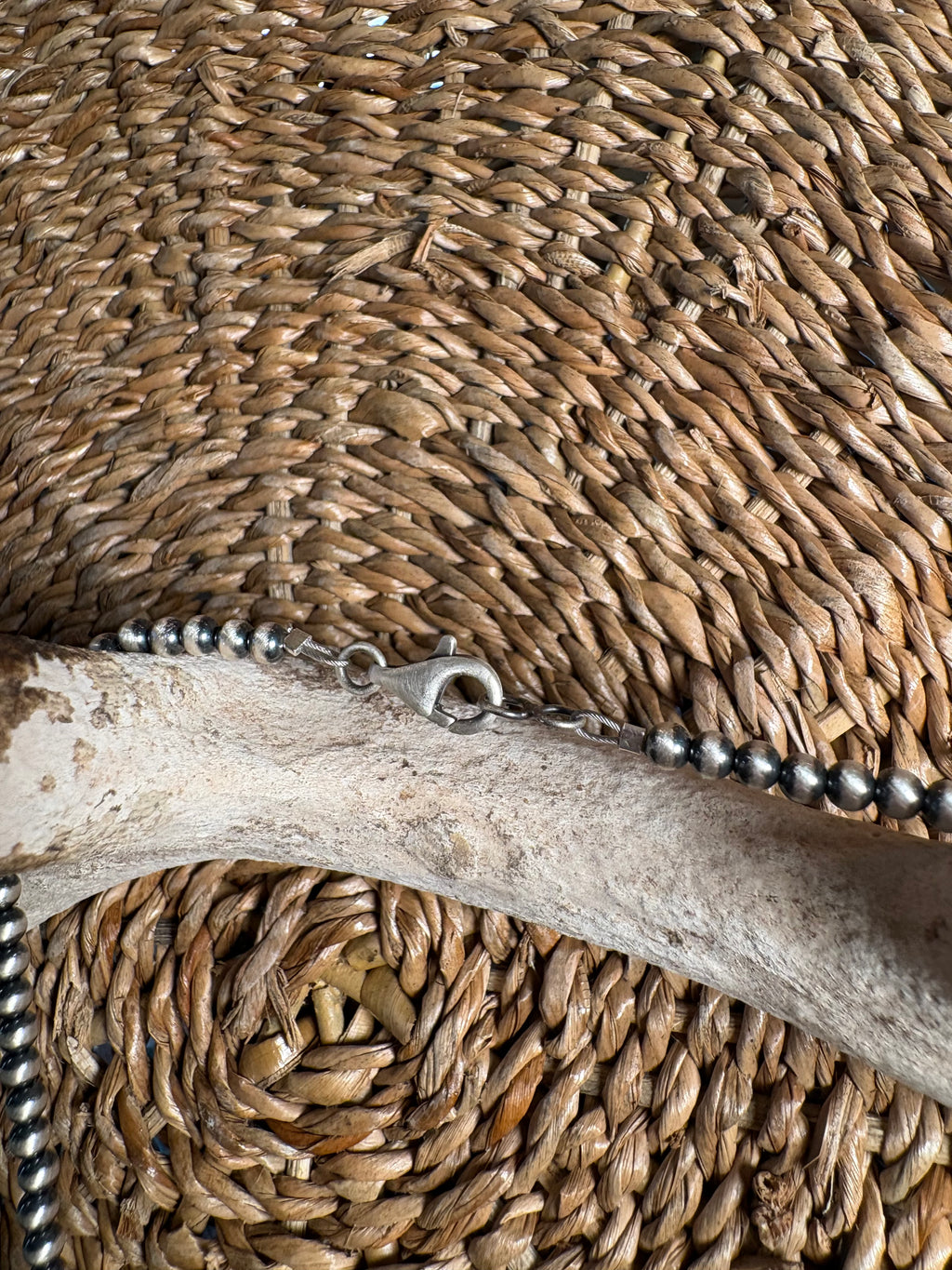 A close-up of a silver beaded chain necklace with a lobster clasp, laid across a natural bone prop on a woven straw mat background.