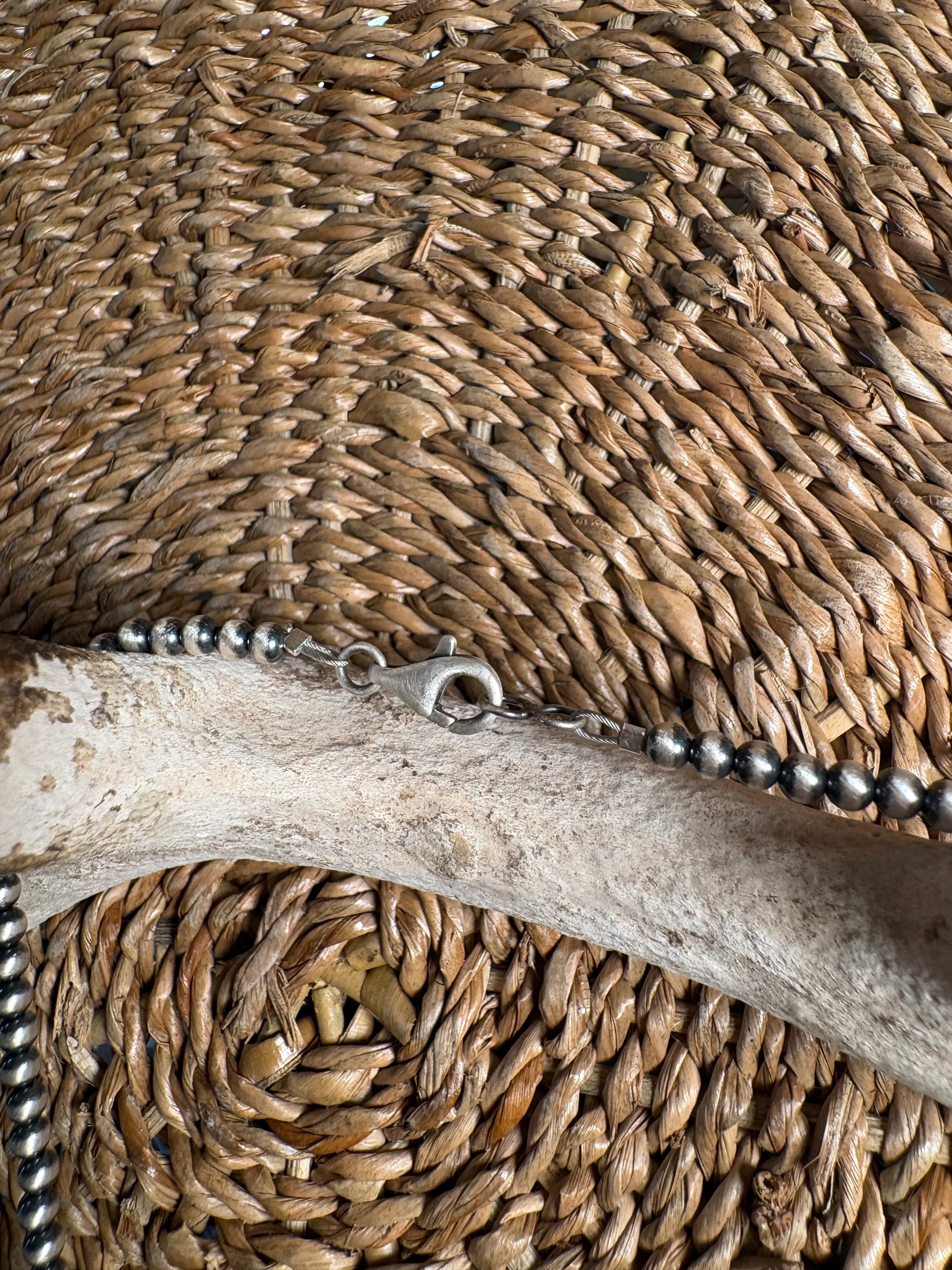 A close-up of a silver beaded chain necklace with a lobster clasp, laid across a natural bone prop on a woven straw mat background.