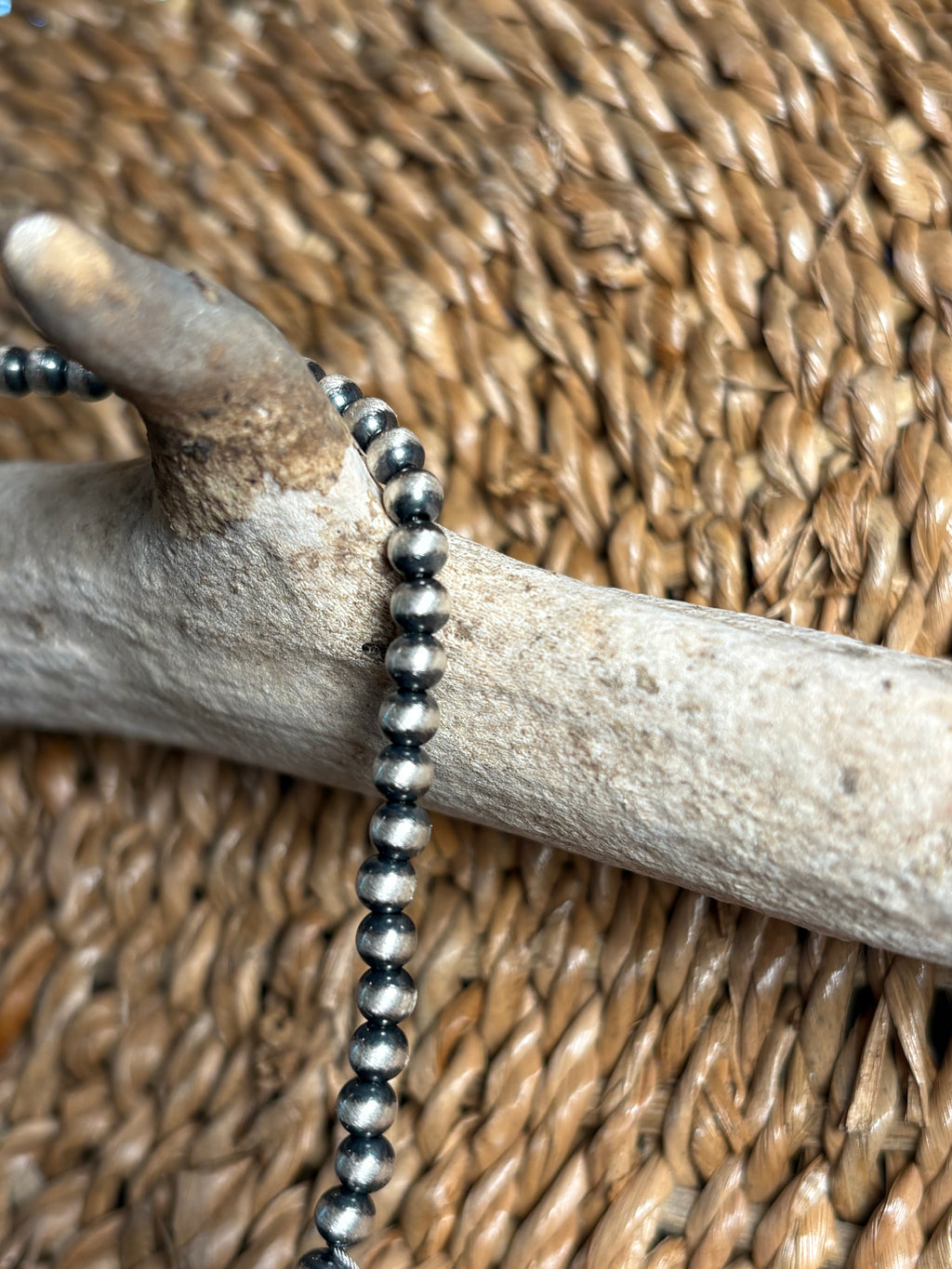 A close-up of a beaded necklace with dark gray and silver metallic round beads draped over a light wood branch, set against a woven tan background.