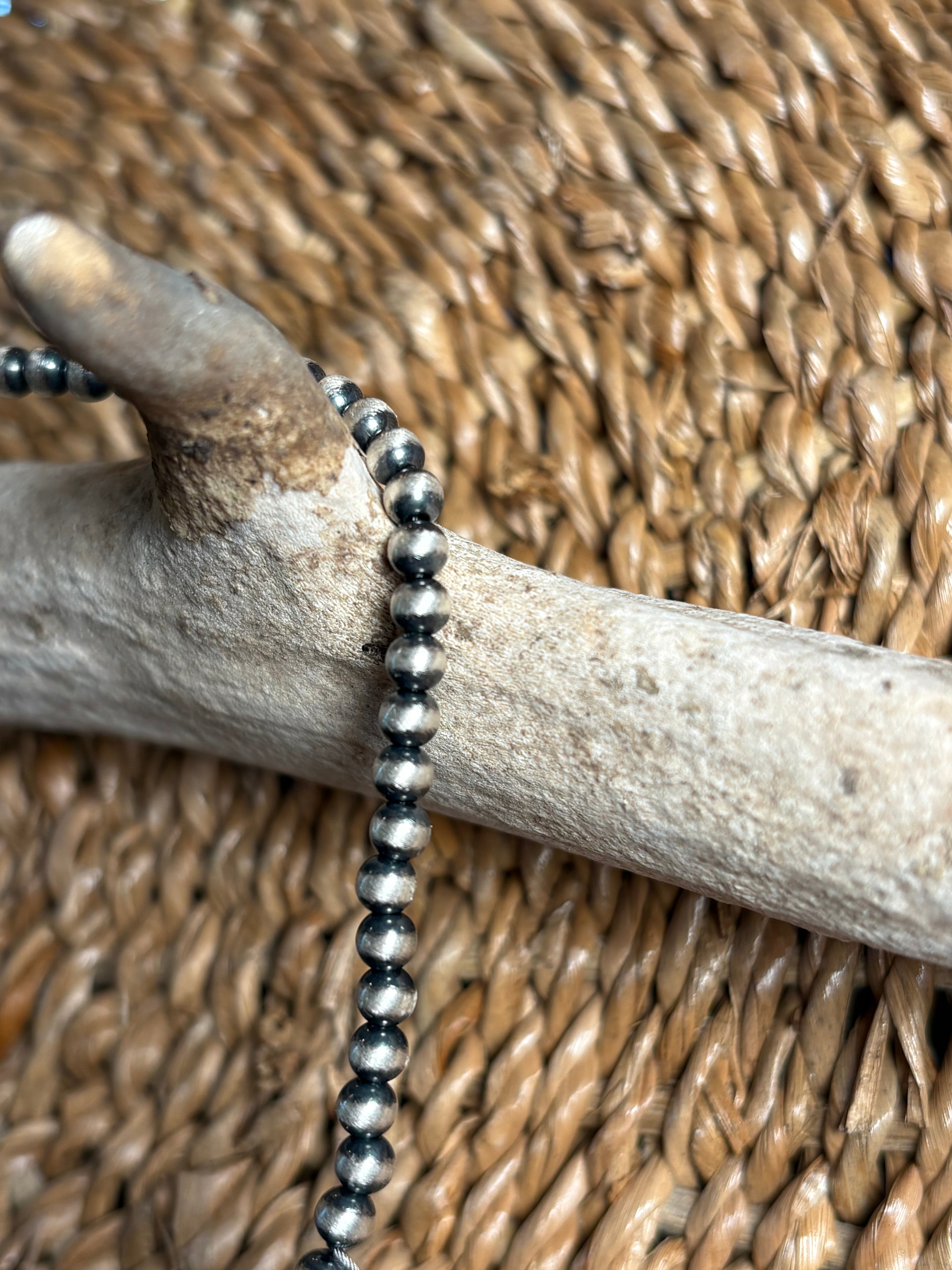 A close-up of a beaded necklace with dark gray and silver metallic round beads draped over a light wood branch, set against a woven tan background.