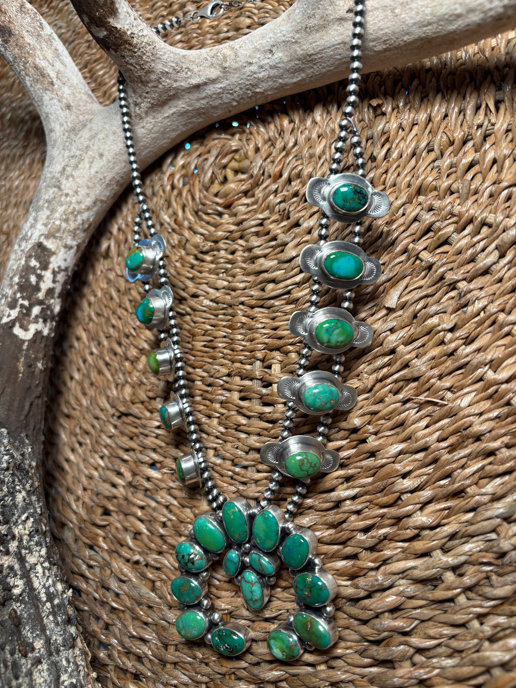A silver beaded necklace with multiple green turquoise cabochon and oval stone settings, displayed draped over a woven brown basket with an antler prop.