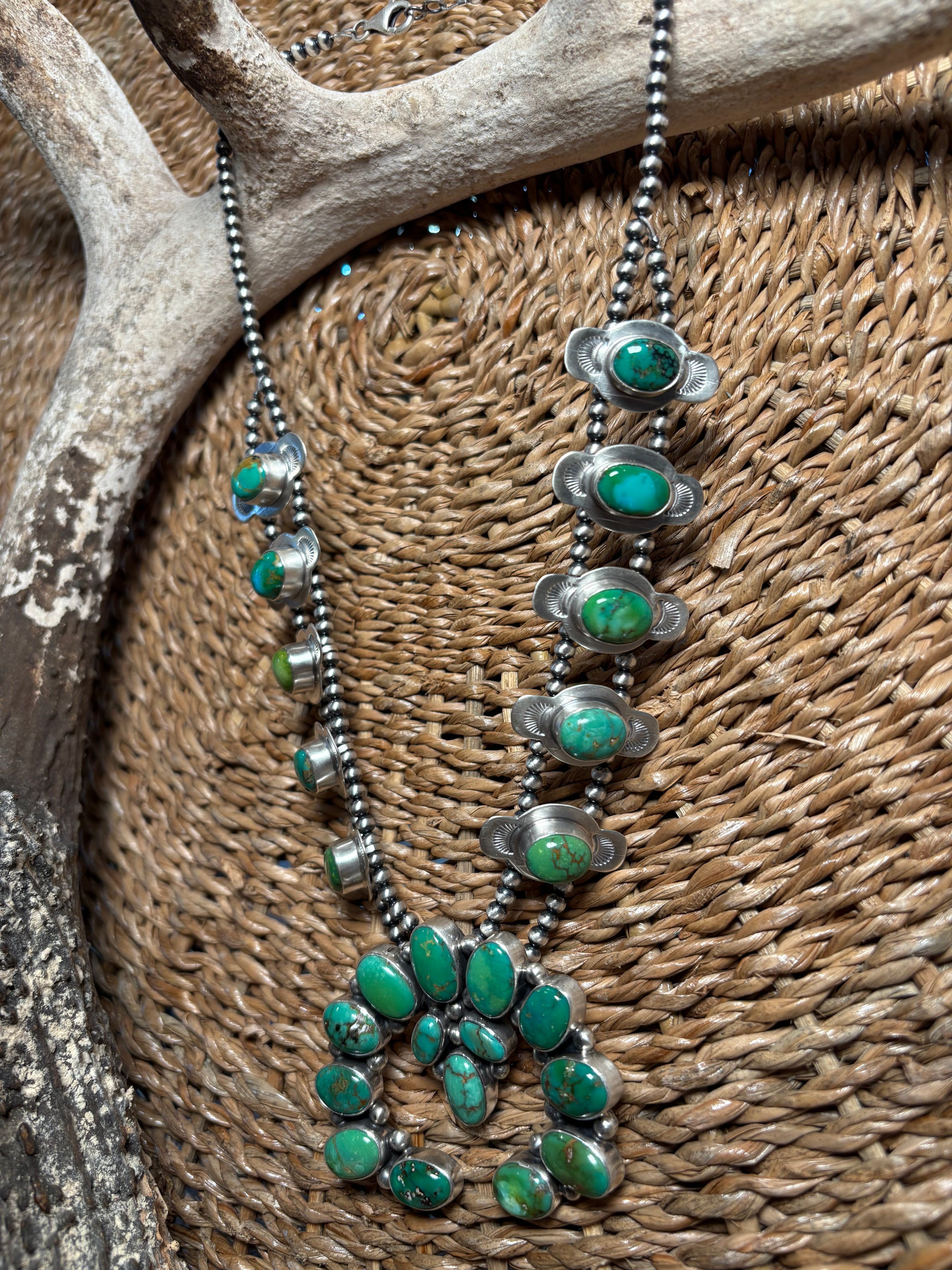 A silver beaded necklace with multiple green turquoise cabochon and oval stone settings, displayed draped over a woven brown basket with an antler prop.