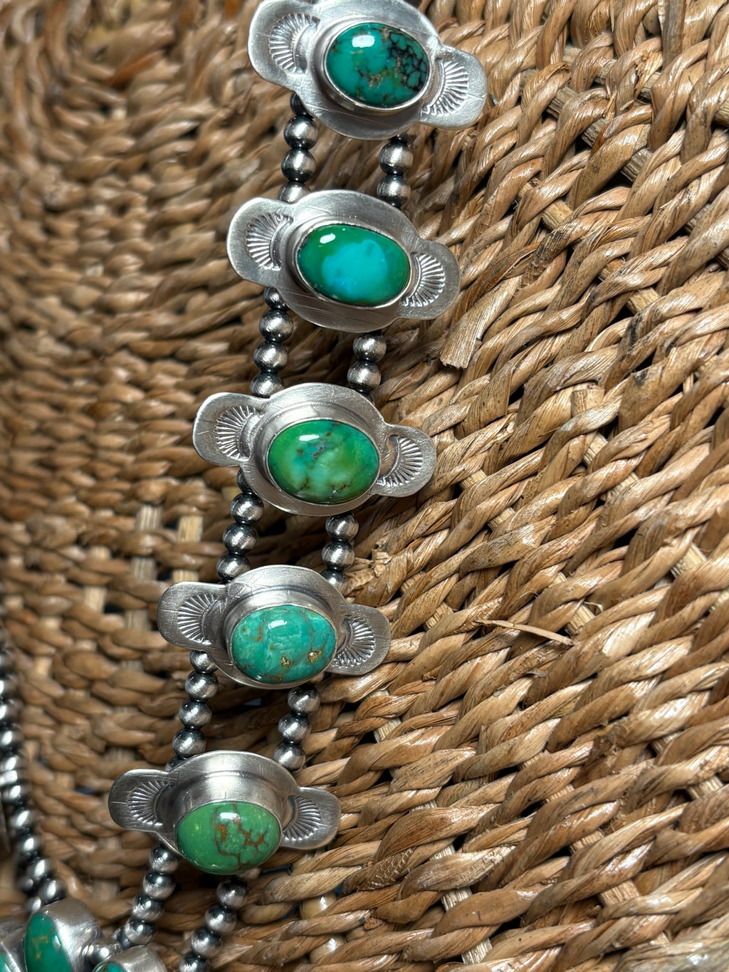 A close-up of a silver beaded necklace with oval turquoise stones, displayed on a woven brown straw surface.