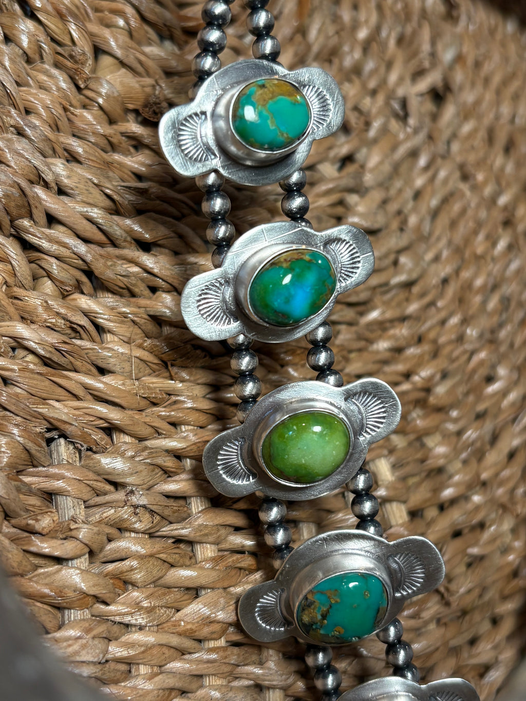 A close-up of a silver beaded necklace with four oval turquoise and green stone settings, resting on a woven brown straw surface.