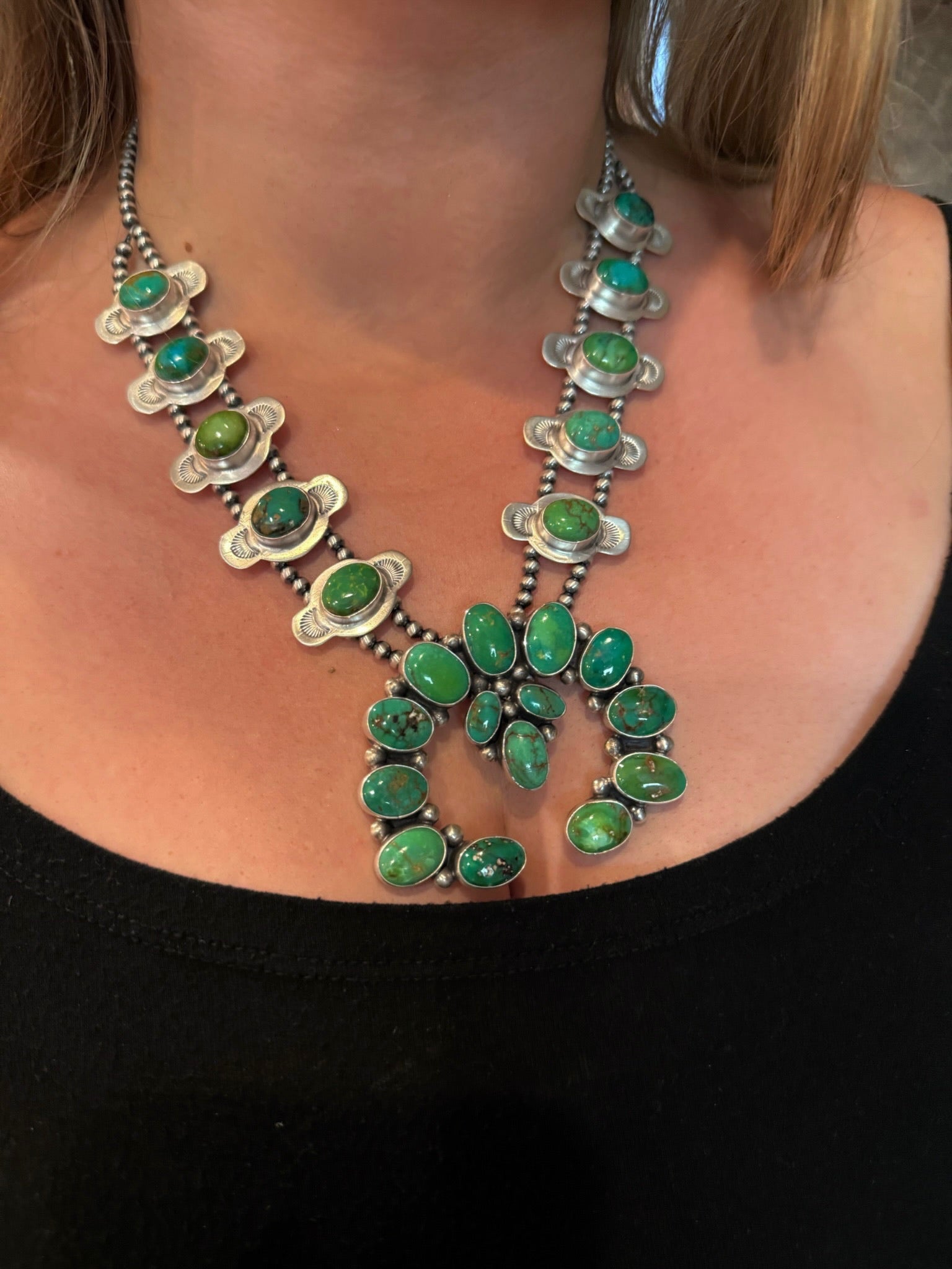 A close-up of a Southwestern-style silver and turquoise necklace worn by a person, featuring multiple green turquoise cabochons set in silver, paired with a black top.