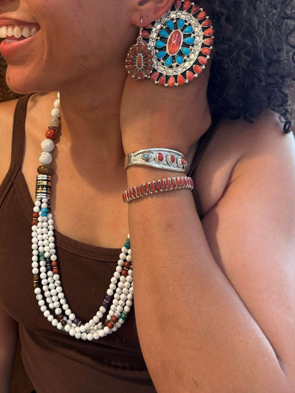 A woman wearing a multi-strand white and turquoise beaded necklace, a matching beaded bracelet, and a large round turquoise and coral earring, shown in a close-up portrait.